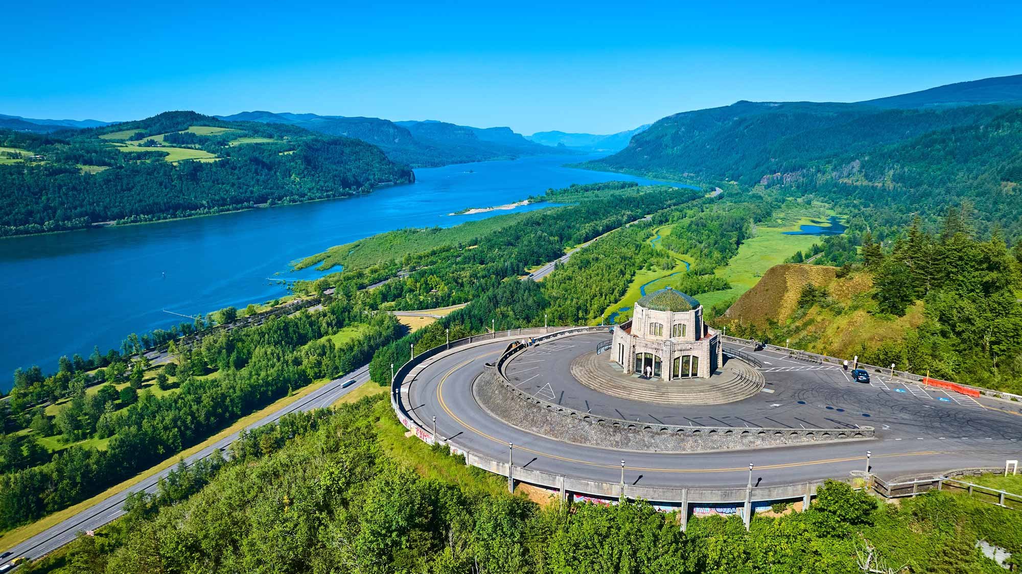 Scenic view of a winding road and a small round building with an observation deck, overlooking a river and lush green landscape with mountains in the background.
