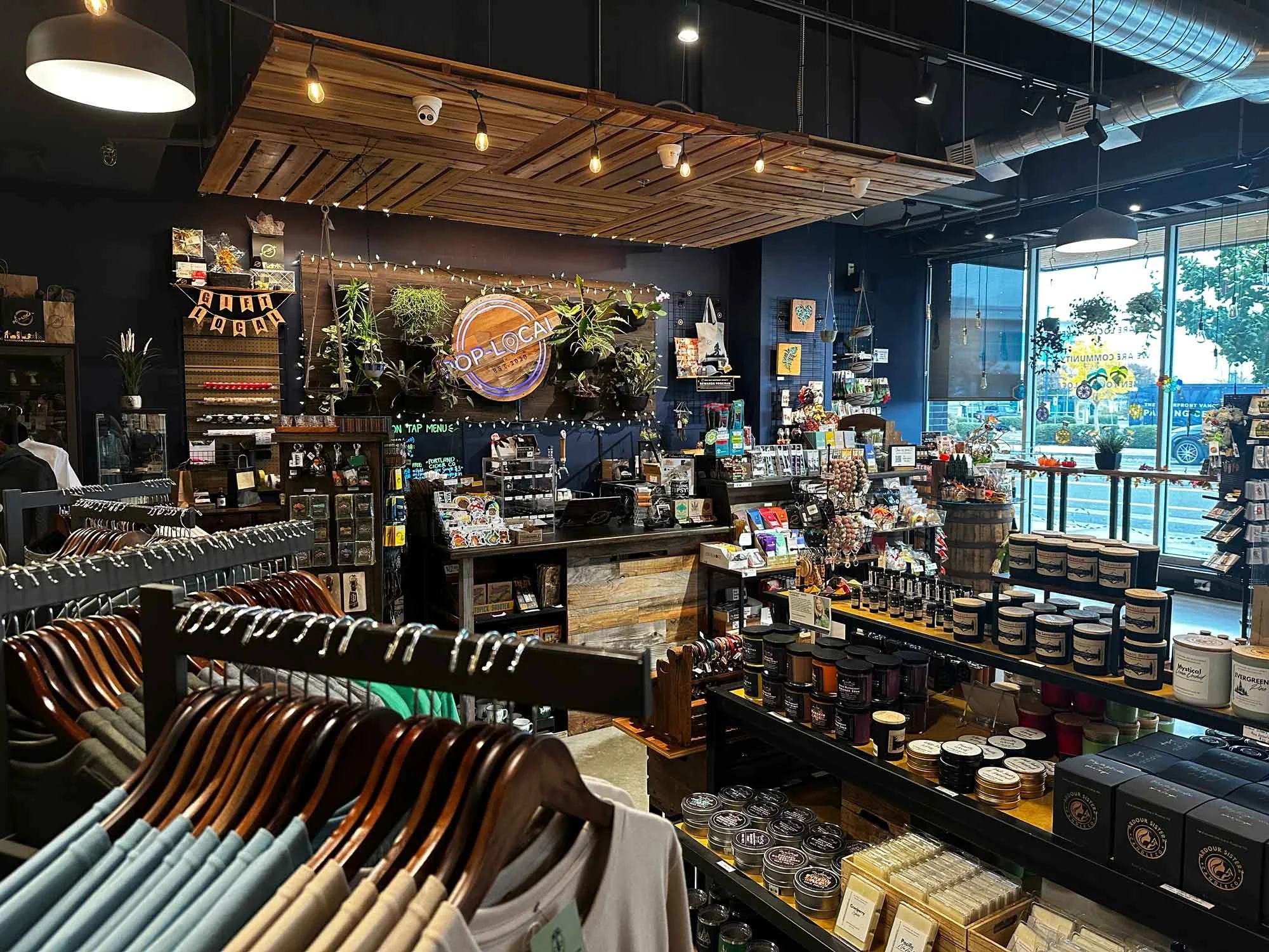 A retail store interior with clothing on hangers in the foreground and shelves with various products including skincare and gift items. The store has a modern industrial design with wooden accents, exposed ductwork, and large windows.