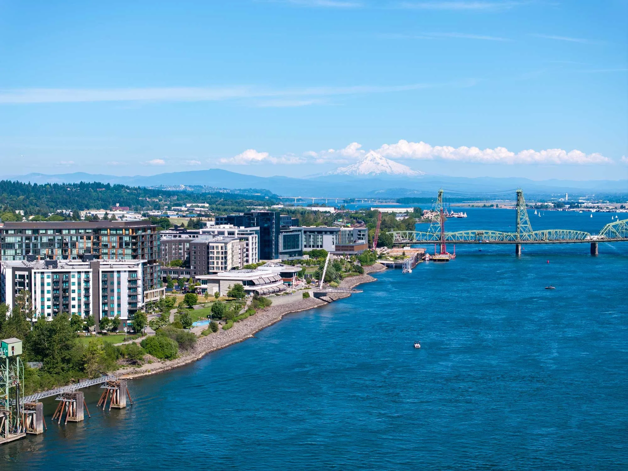 Cityscape with modern buildings along a river, a bridge, forests, and mountains including Mount Rainier in the background under a blue sky with some clouds.