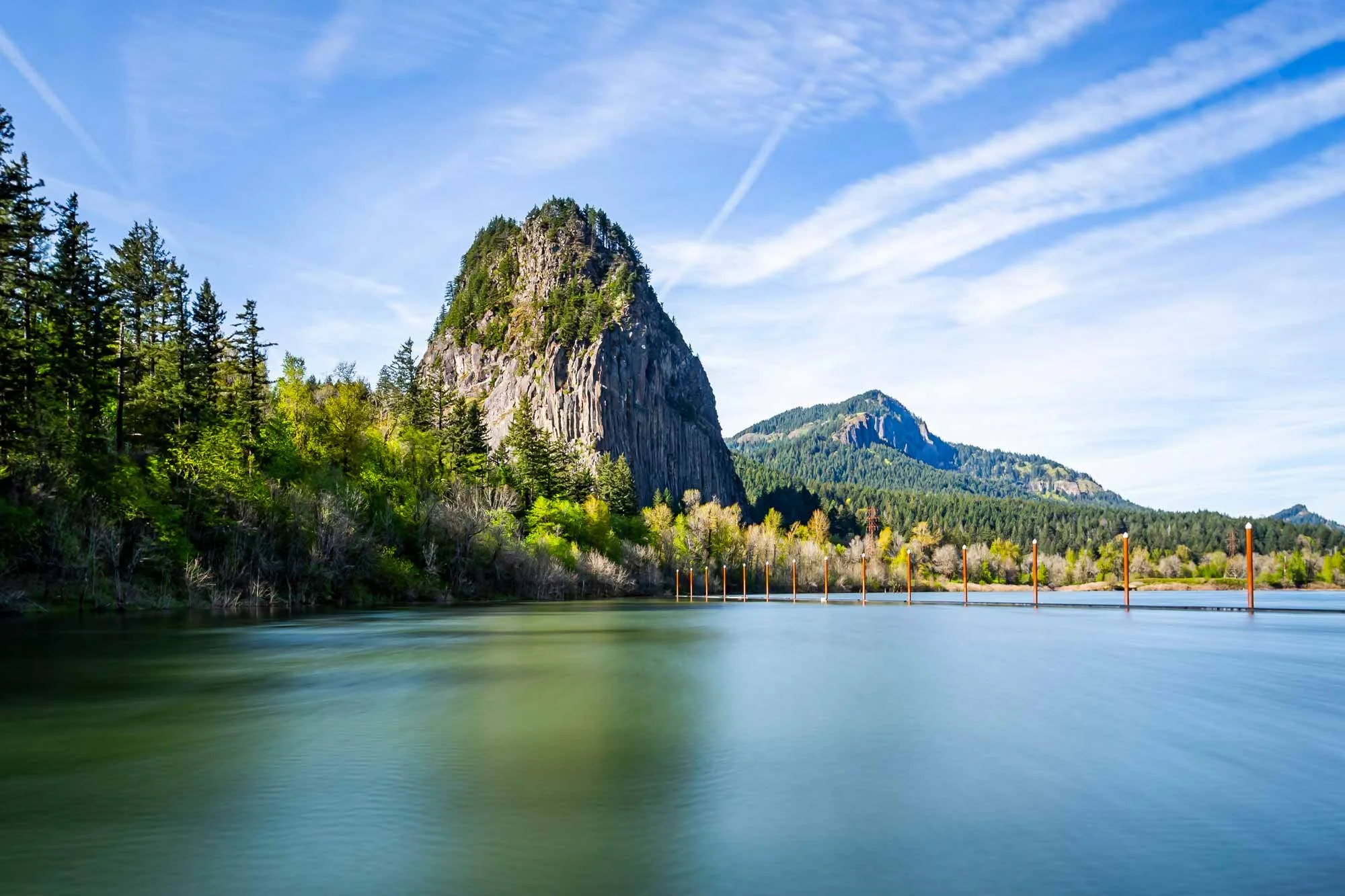 A scenic landscape of a mountain with greenery, a river in the foreground, and a partly cloudy sky with jet trails.