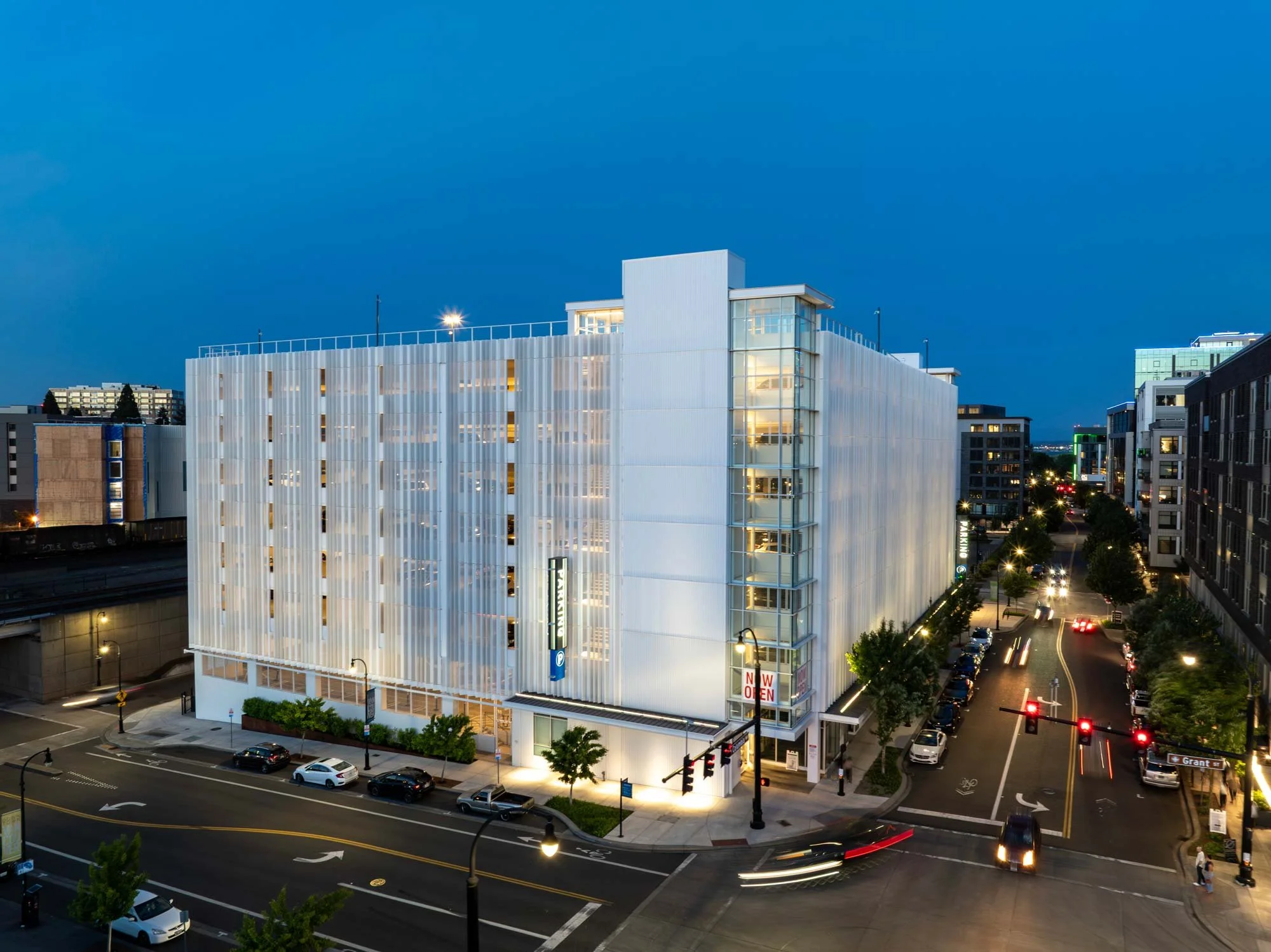 Nighttime cityscape featuring a modern white building with illuminated windows, parking lot, cars on the streets, and other buildings in the background under a dark blue sky.