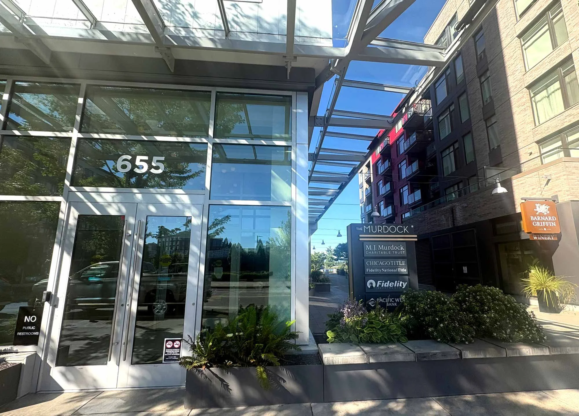 The entrance to a modern building with glass doors and the number 655 displayed on the window. There are signs outside indicating it houses businesses such as M.J. Murdock Charitable Trust, Chicago Title, Fidelity National Title, and Pacific Western Bank. The building has a contemporary design with glass, metal framework, and some greenery in the foreground.