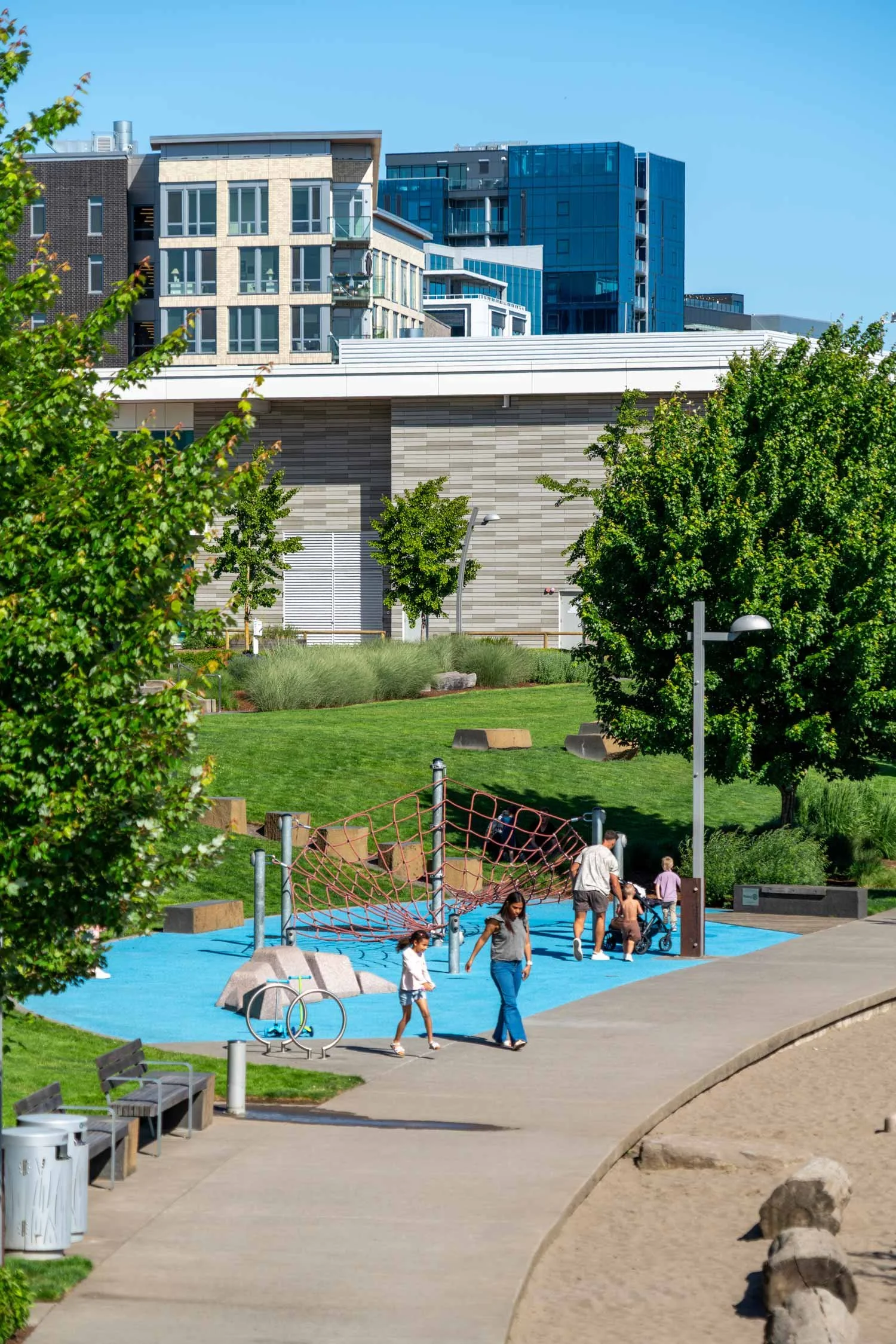 A playground with children and adults, featuring a red net climbing structure on a blue rubber surface, surrounded by green trees and park benches, with modern buildings in the background.