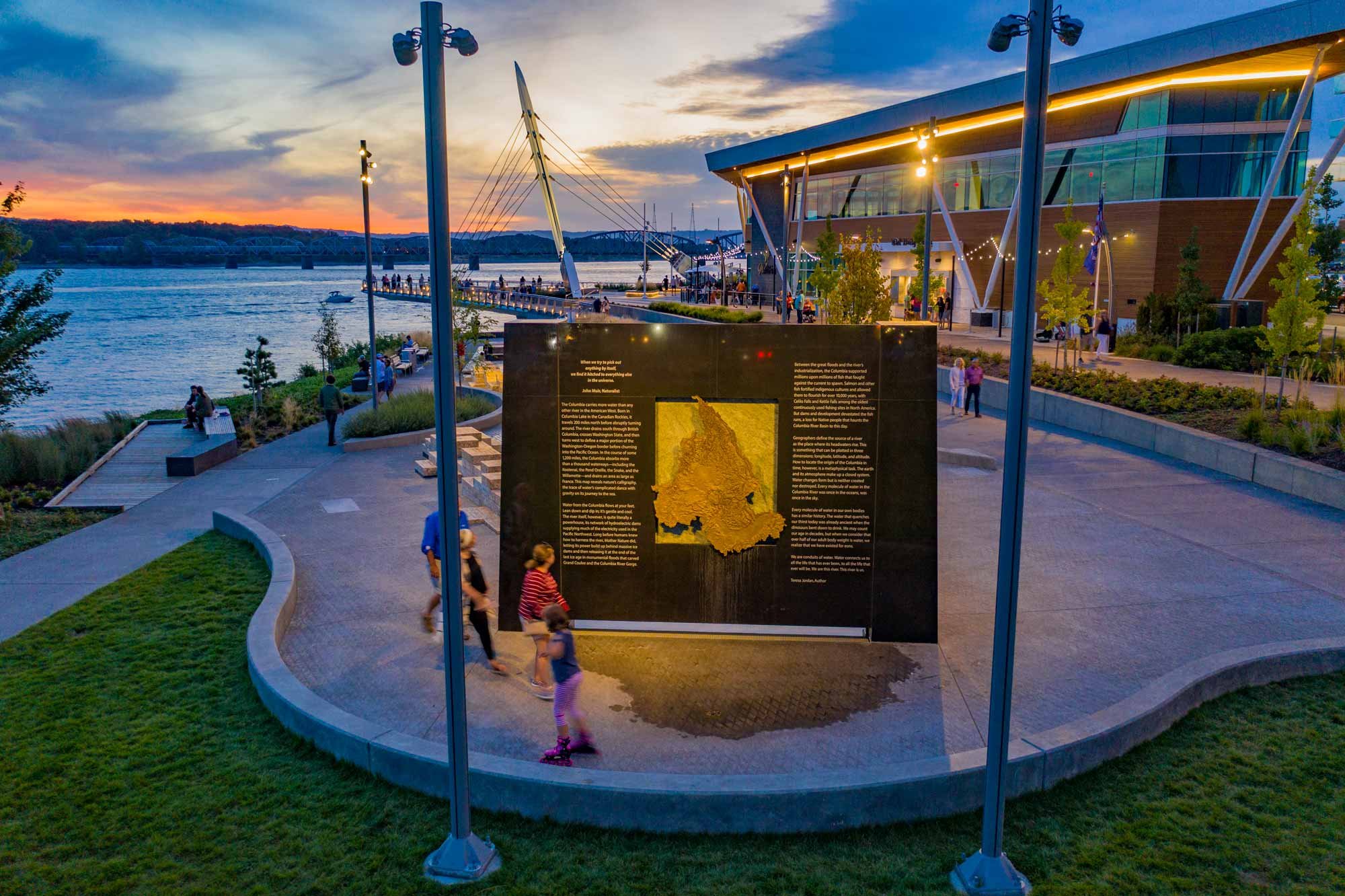 People walking and sitting at a scenic overlook near a body of water at sunset, with a modern building and lampposts in the background.