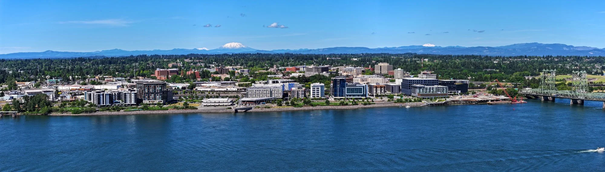 A city skyline along a river with modern buildings, a bridge, and mountains with snow in the background, under a clear blue sky.