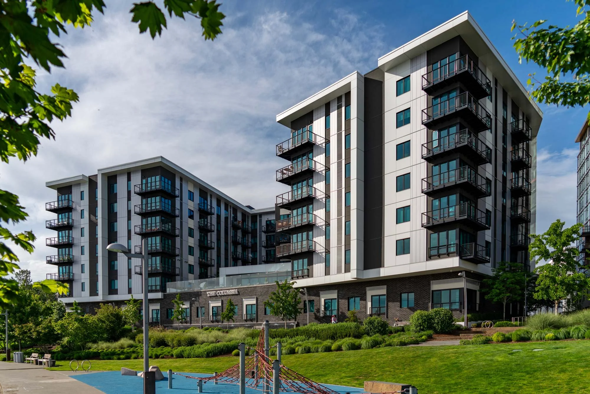 Modern multi-story apartment buildings with balconies in an urban park setting, with trees, greenery, benches, and a playground area in the foreground.