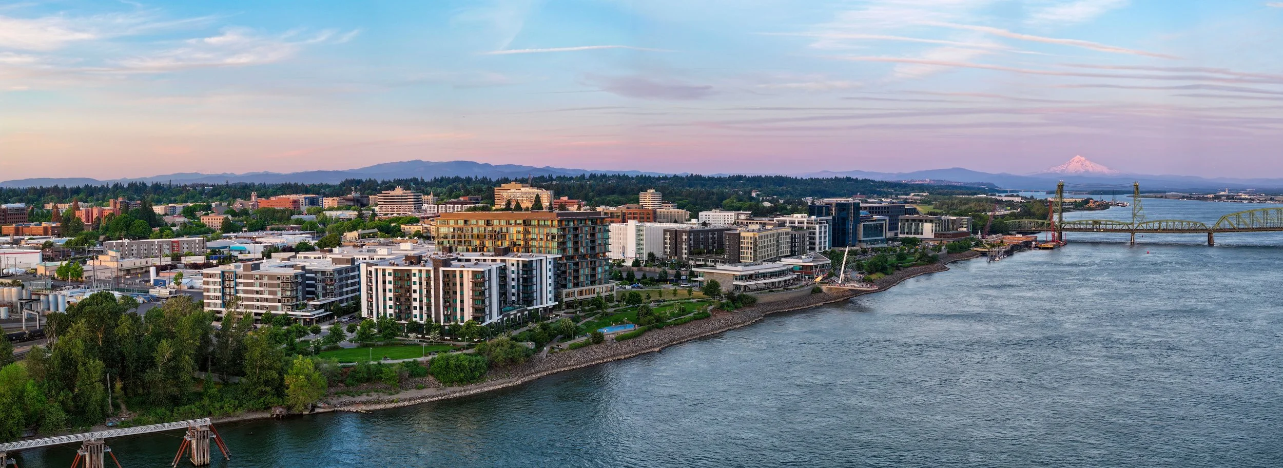 A panoramic view of a city by the river with modern buildings, a bridge, and a mountain in the background during sunset.