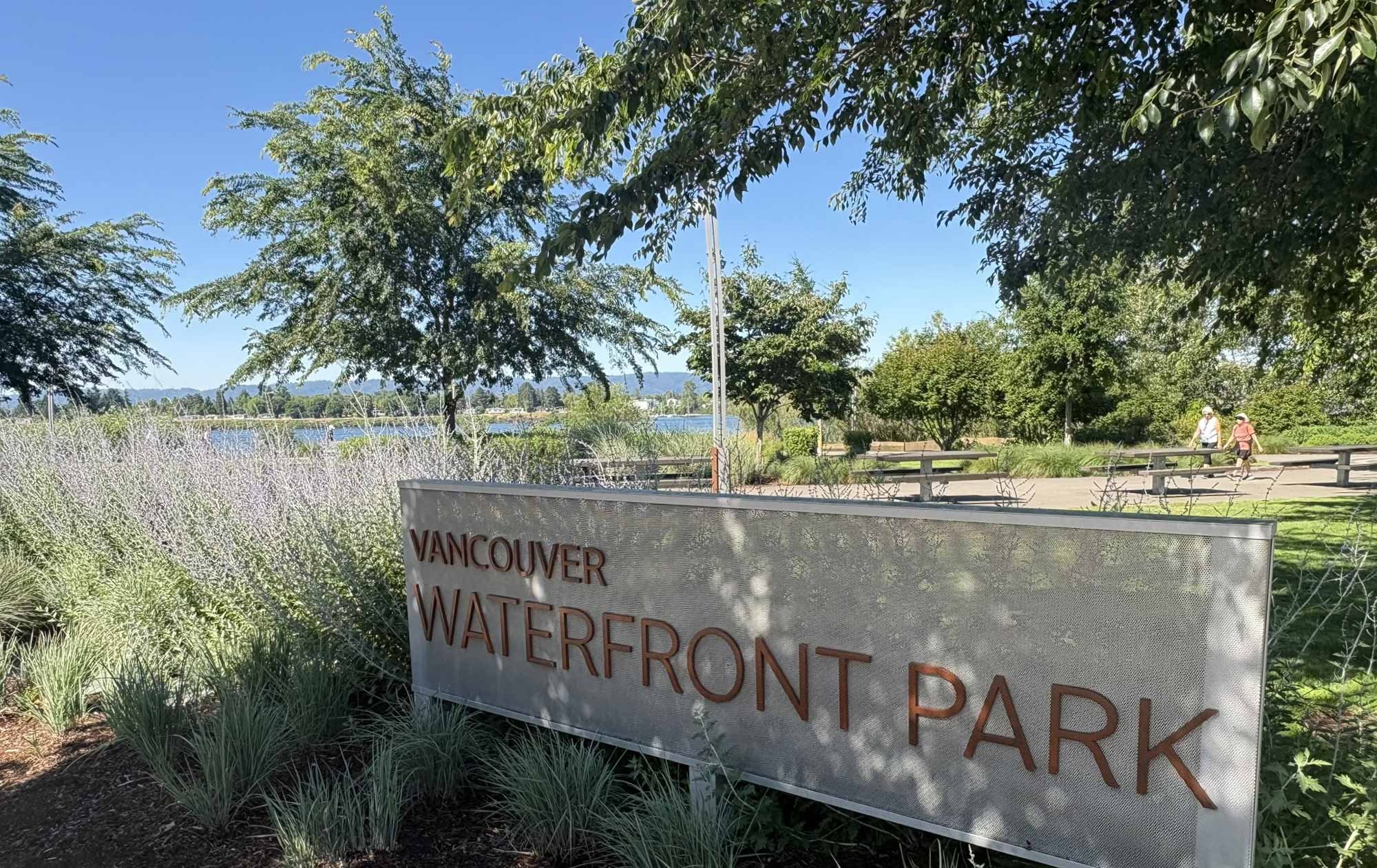A sign reading 'Vancouver Waterfront Park' in front of trees, greenery, and a view of the water with a bridge and two people walking.