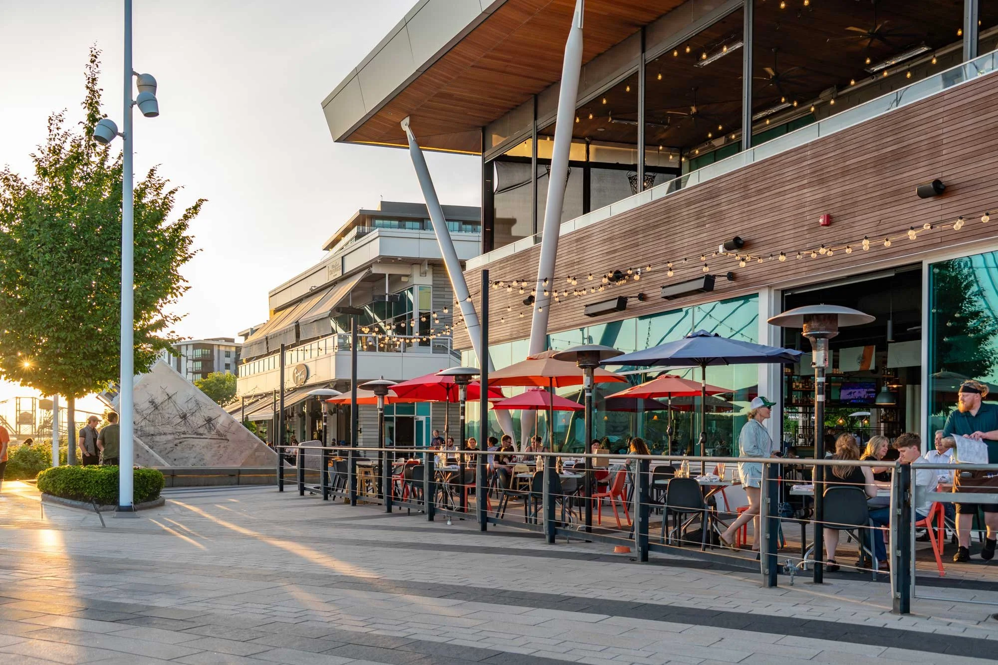 People dining and socializing outdoors at a modern shopping center with a glass and wooden building, string lights, umbrellas, and trees, during sunset.