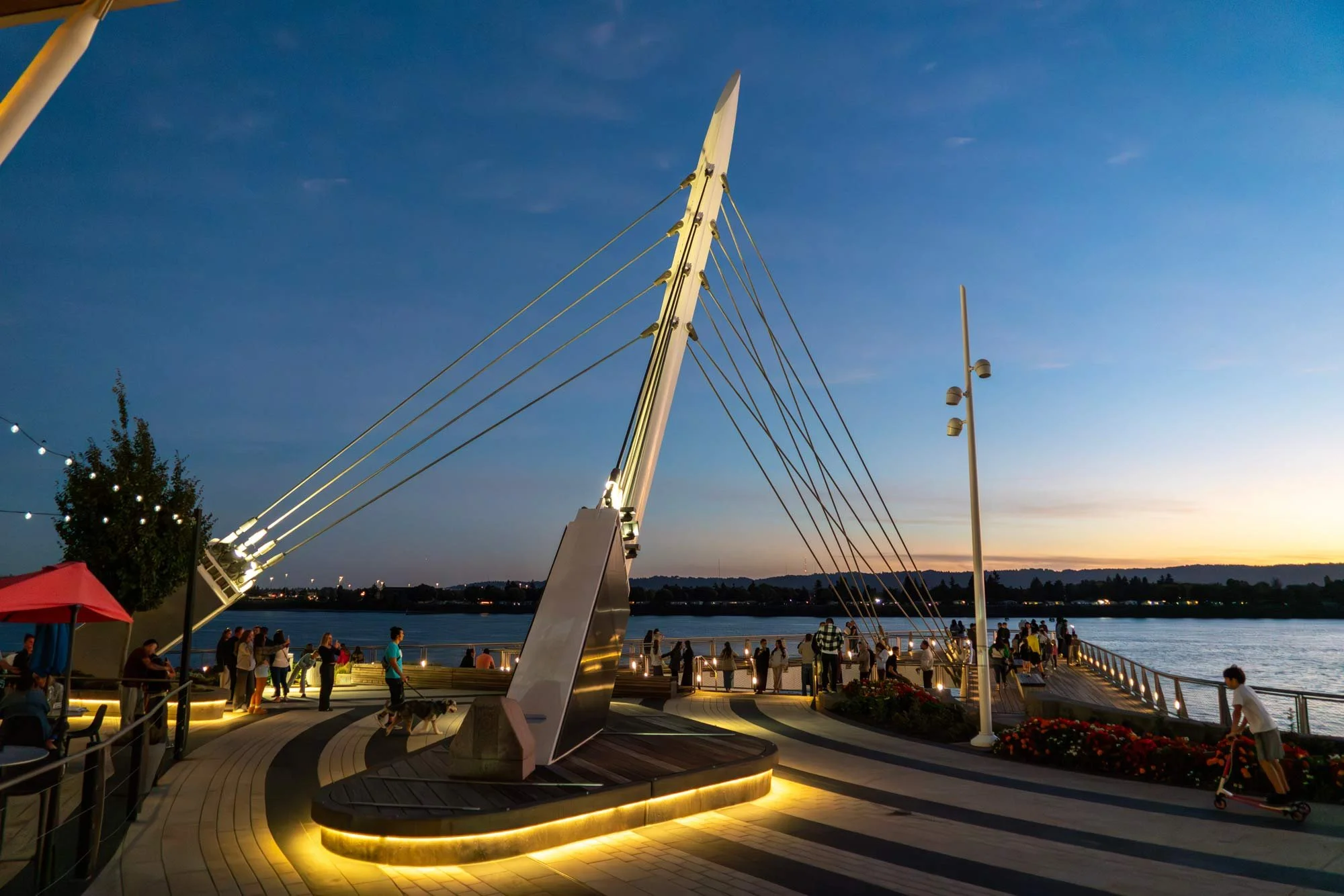 View of a modern suspension bridge with electric lighting, a river, and a crowd of people on a wooden pier during dusk, with a clear sky and distant hills.