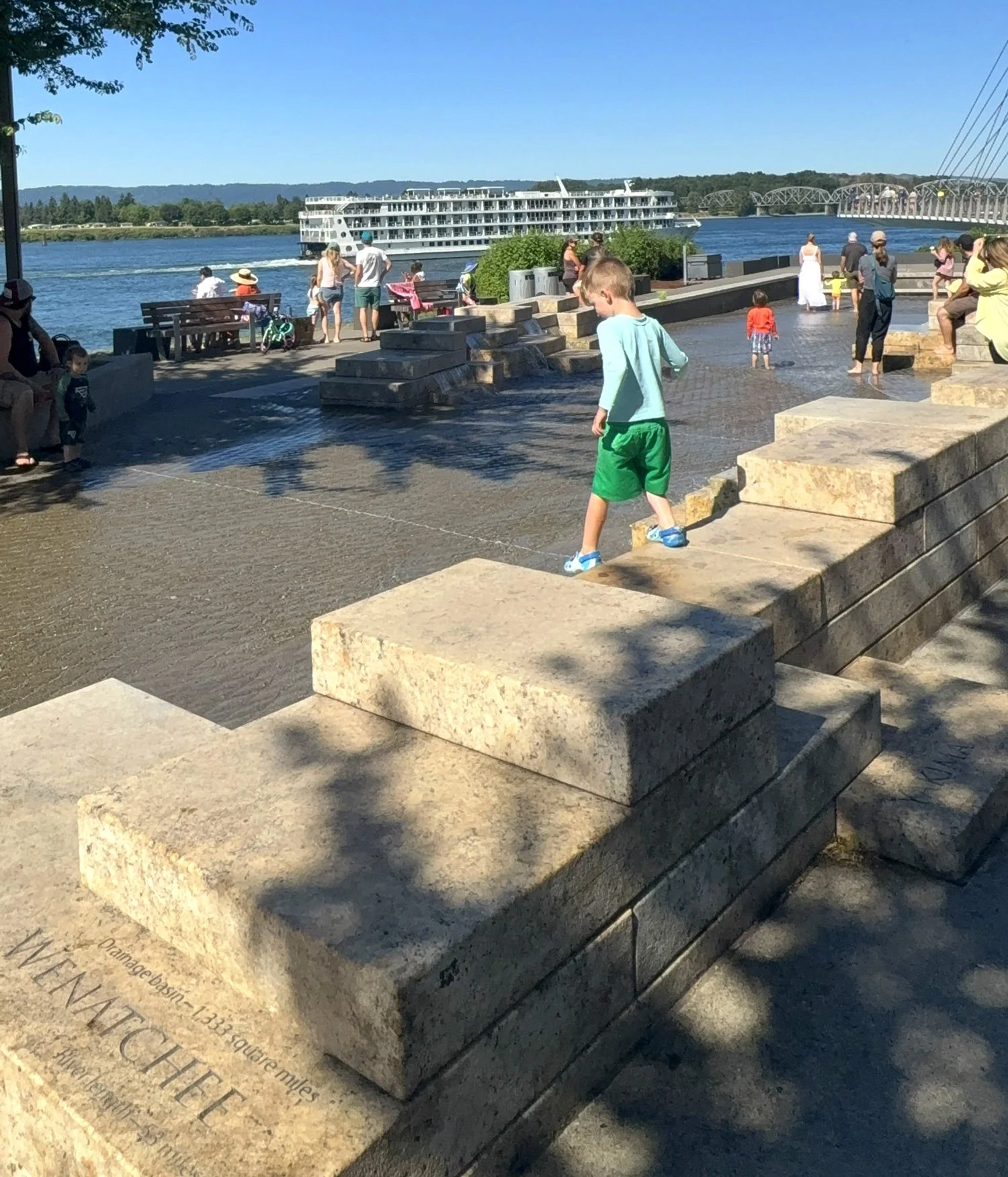 People gather near a river under a clear blue sky, with a large river cruise ship passing by and a bridge in the background. Some children are playing in water, and others sit on benches or walk along the riverbank.
