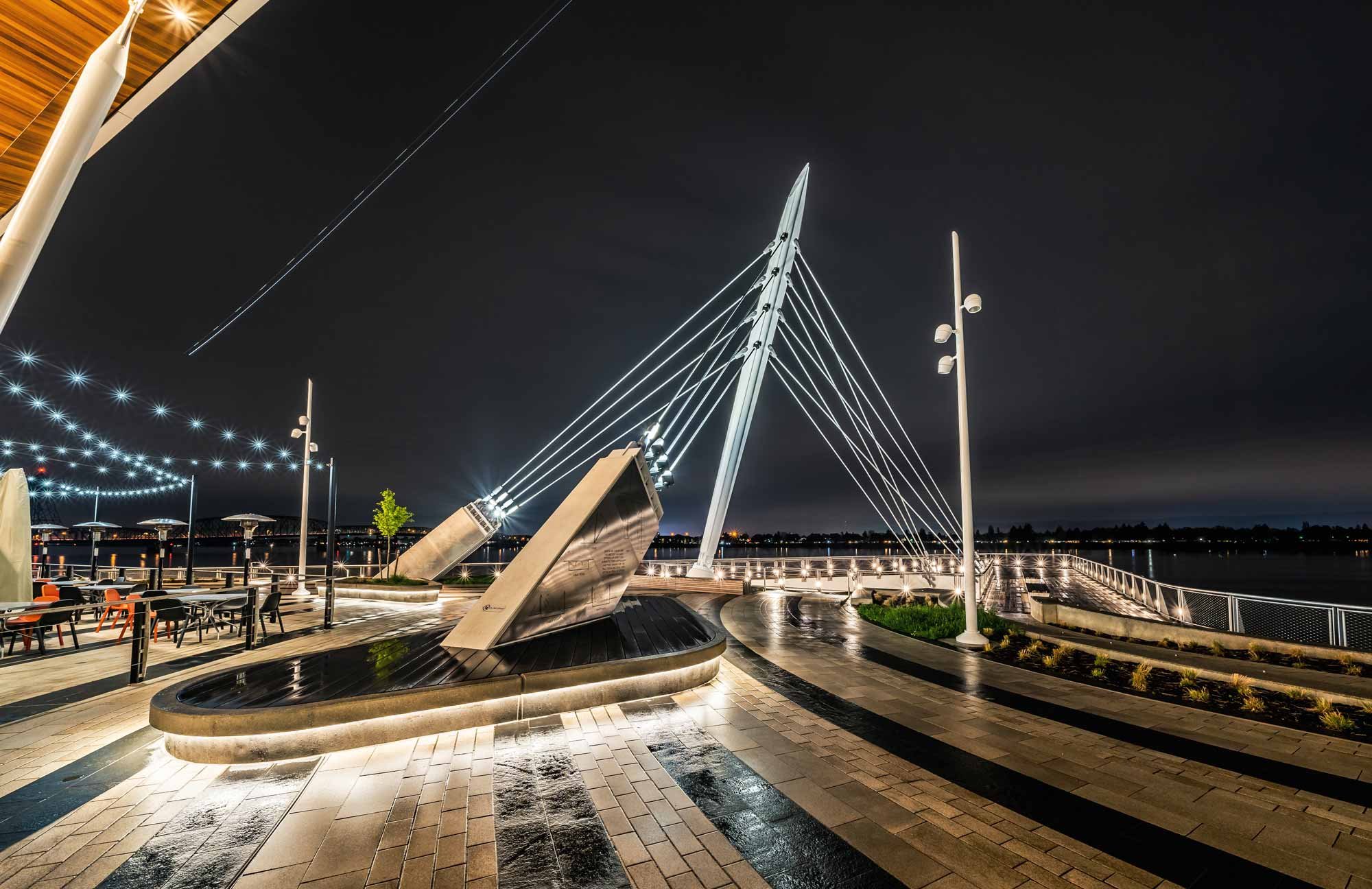 Nighttime view of a modern pedestrian bridge with illuminated cables and a central steel pylon, over a river in a city.