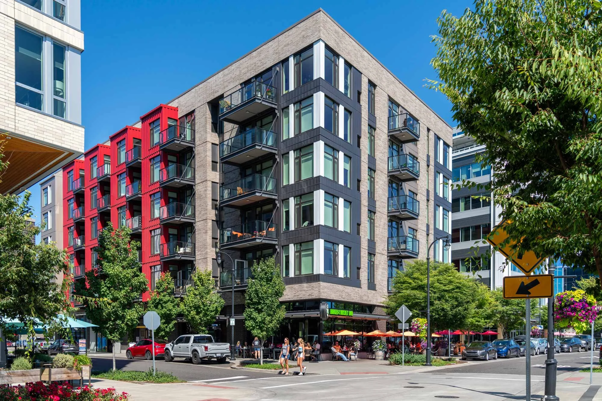 City street scene with a modern multi-story apartment building featuring glass balconies and red architectural accents. Pedestrians walking, cars parked and driving, outdoor seating with umbrellas, lush green trees, and clear blue sky.