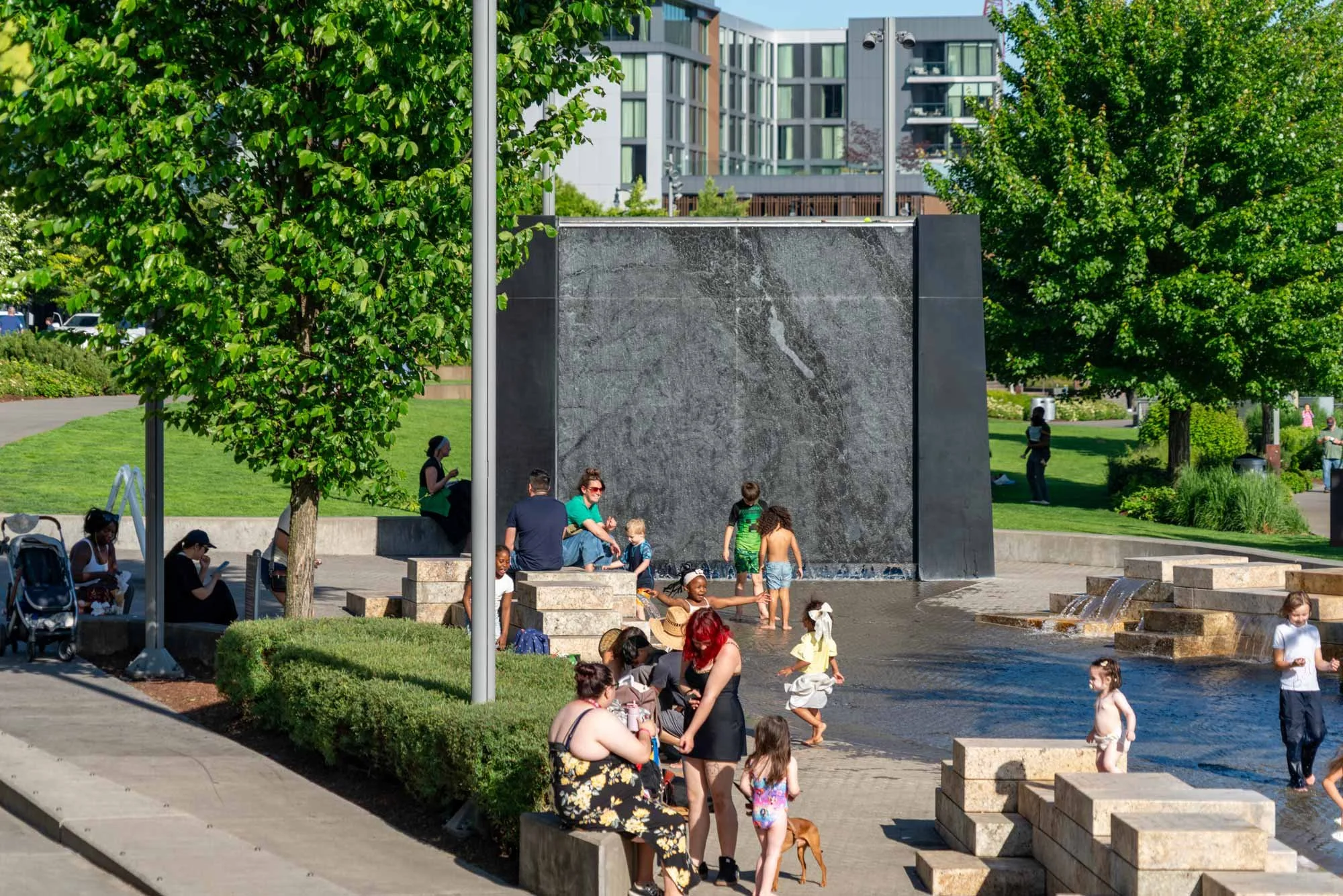 Children and adults playing and relaxing by a water feature in a city park on a sunny day, with a modern apartment building in the background.