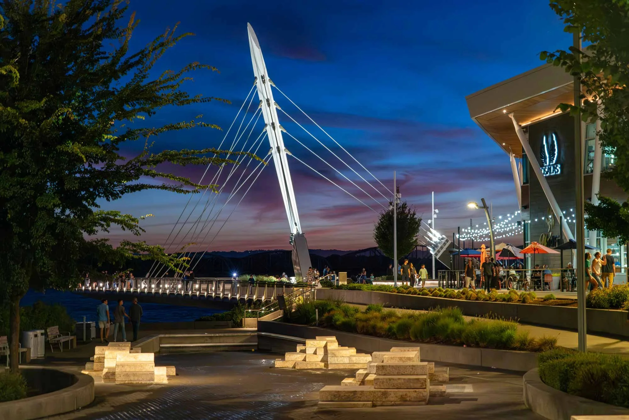 A lively outdoor area of The Waterfront Vancouver park at dusk with modern architecture, a large white bridge sculpture, string lights, and people walking and socializing.