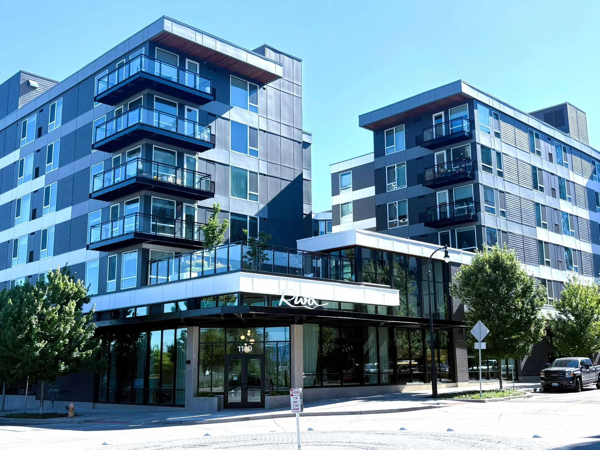 Modern multi-story apartment building with glass balconies and exterior, surrounded by trees and parked cars under a clear blue sky.