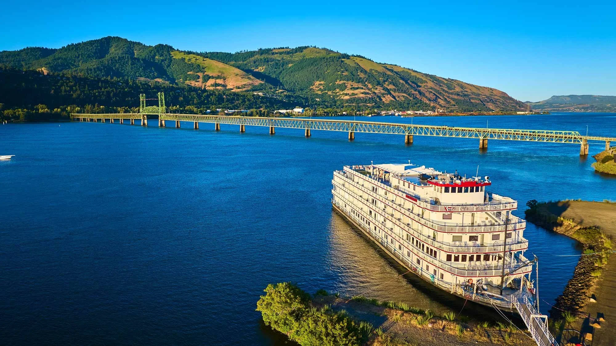 A large cruise ship docked by the river with a bridge and green hills in the background.