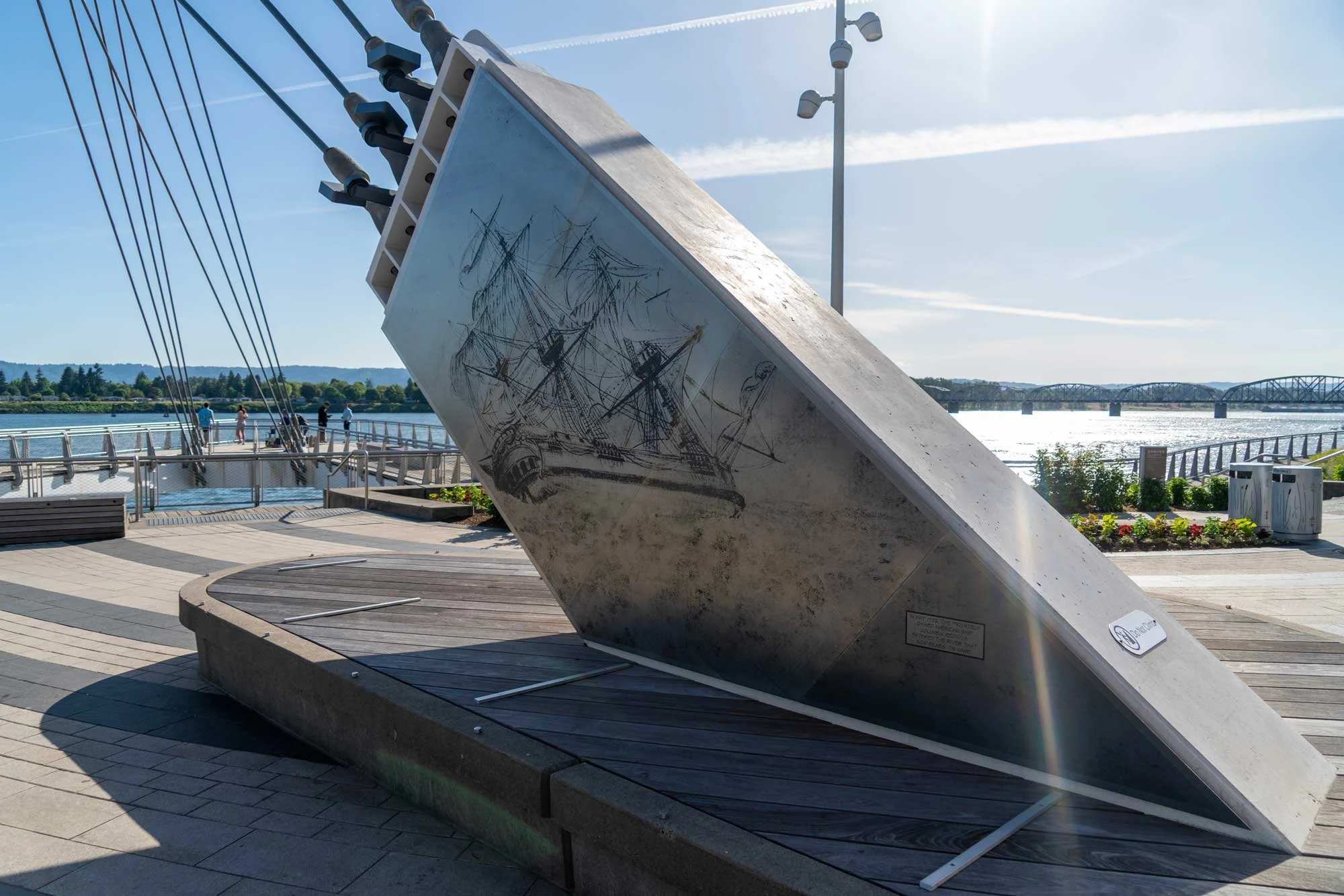 A large outdoor sculpture of a ship's bow with sketch-like drawings of ships, situated on a circular platform by a waterfront. In the background, there are people walking and a bridge over the water under a clear sky with some contrails.