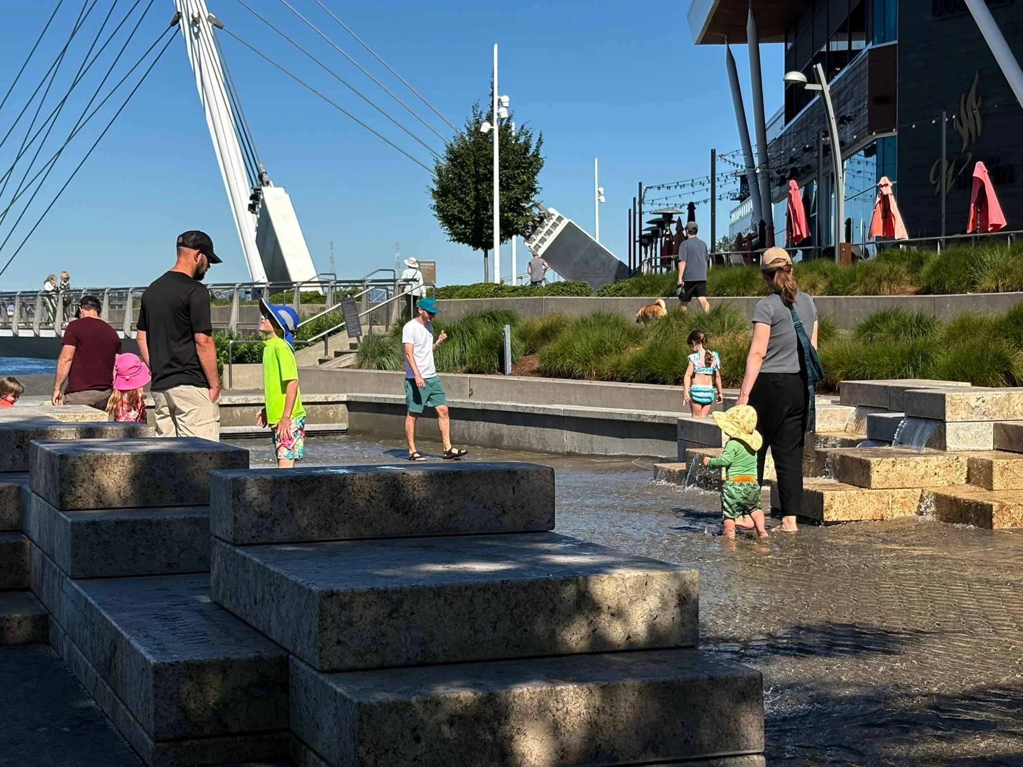 A diverse group of people, including children and adults, are enjoying a sunny day near a water feature with tiered concrete platforms for children to play and splash water. In the background, there is a modern building with large glass windows, red umbrellas on a patio, and a large sailboat mast with rigging, suggesting a waterfront setting.