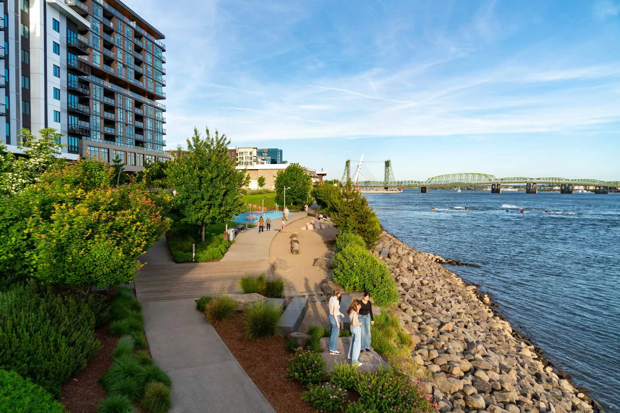 A riverside park with a walking path, trees, and bushes, along a rocky shoreline, with people walking and enjoying the view of a bridge and cityscape across the river.