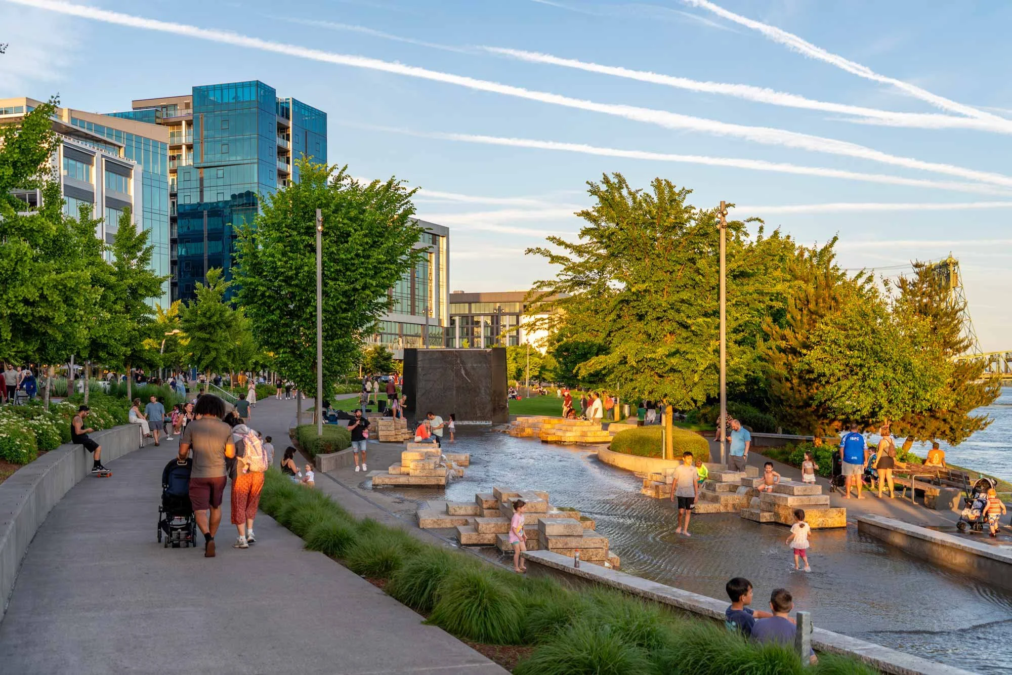 People walking and socializing along a riverside park with trees, a water feature, and modern buildings in the background during what appears to be late afternoon or early evening.