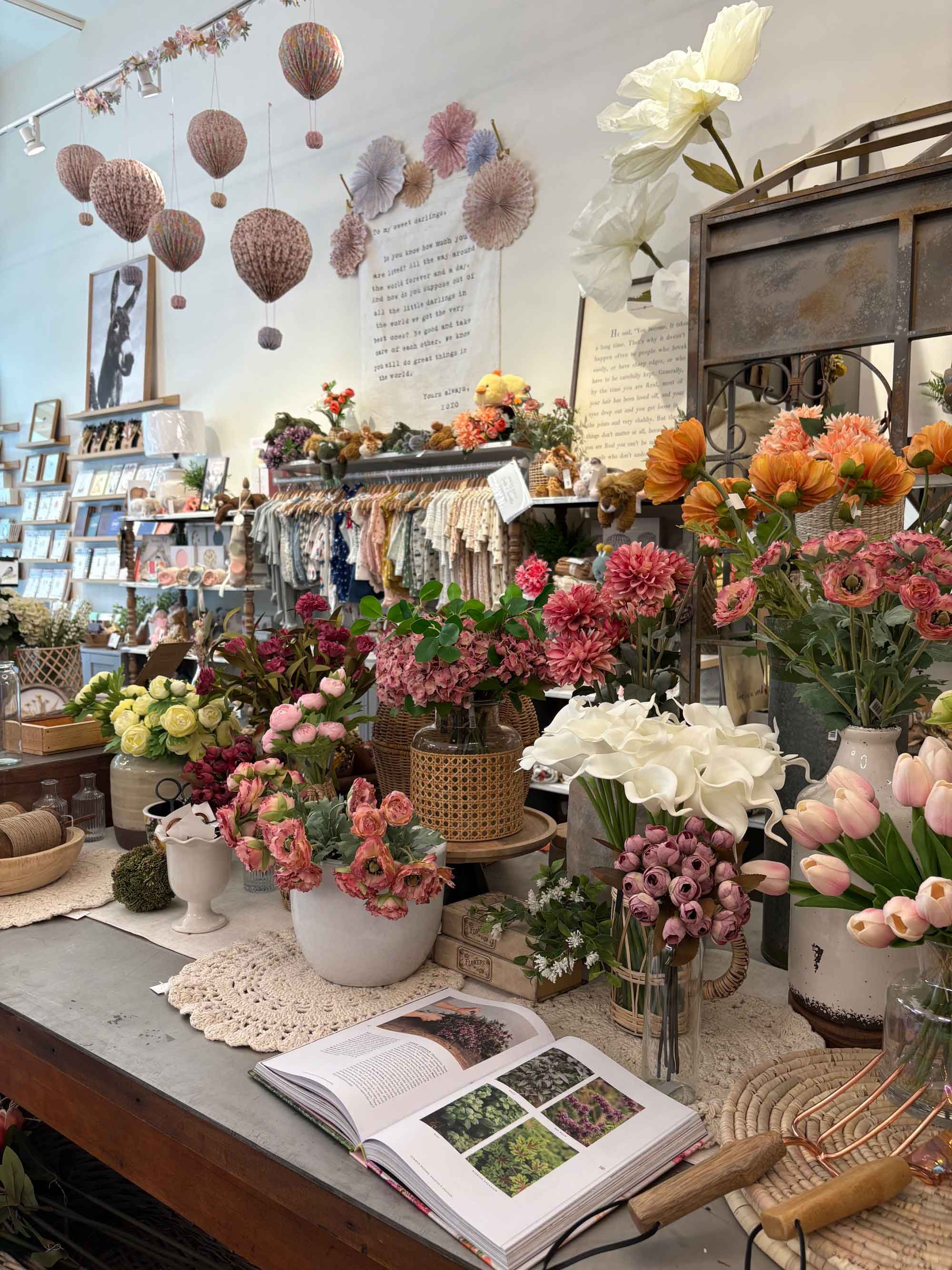Inside a flower shop with various potted and cut flowers, including pink roses, white calla lilies, and peach blooms, displayed on a wooden table with an open book showing plant images. Decorative pink and purple hanging paper lanterns and wall art are visible in the background.