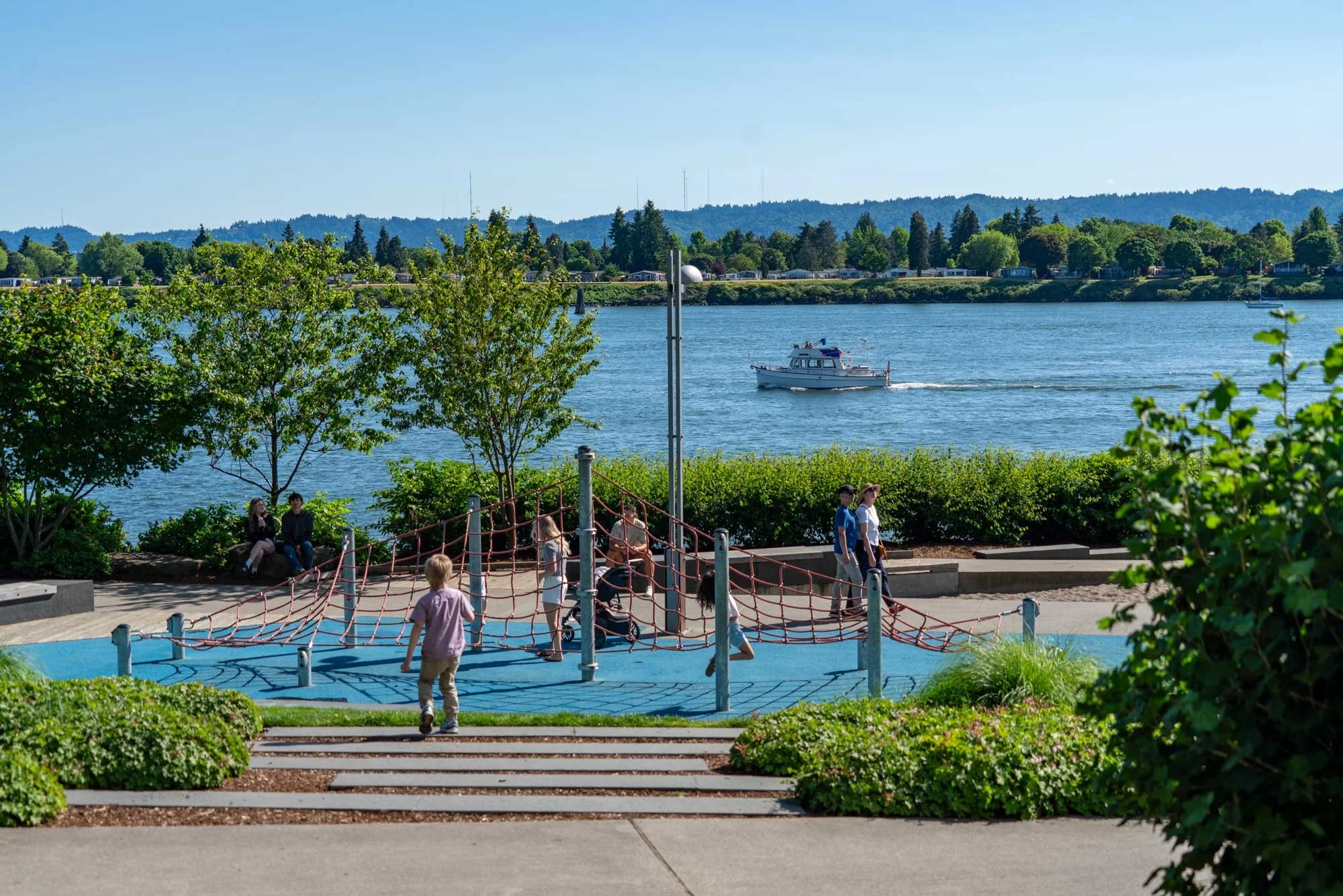 Children playing on a playground near a body of water with a boat, trees, and hills in the background on a sunny day.