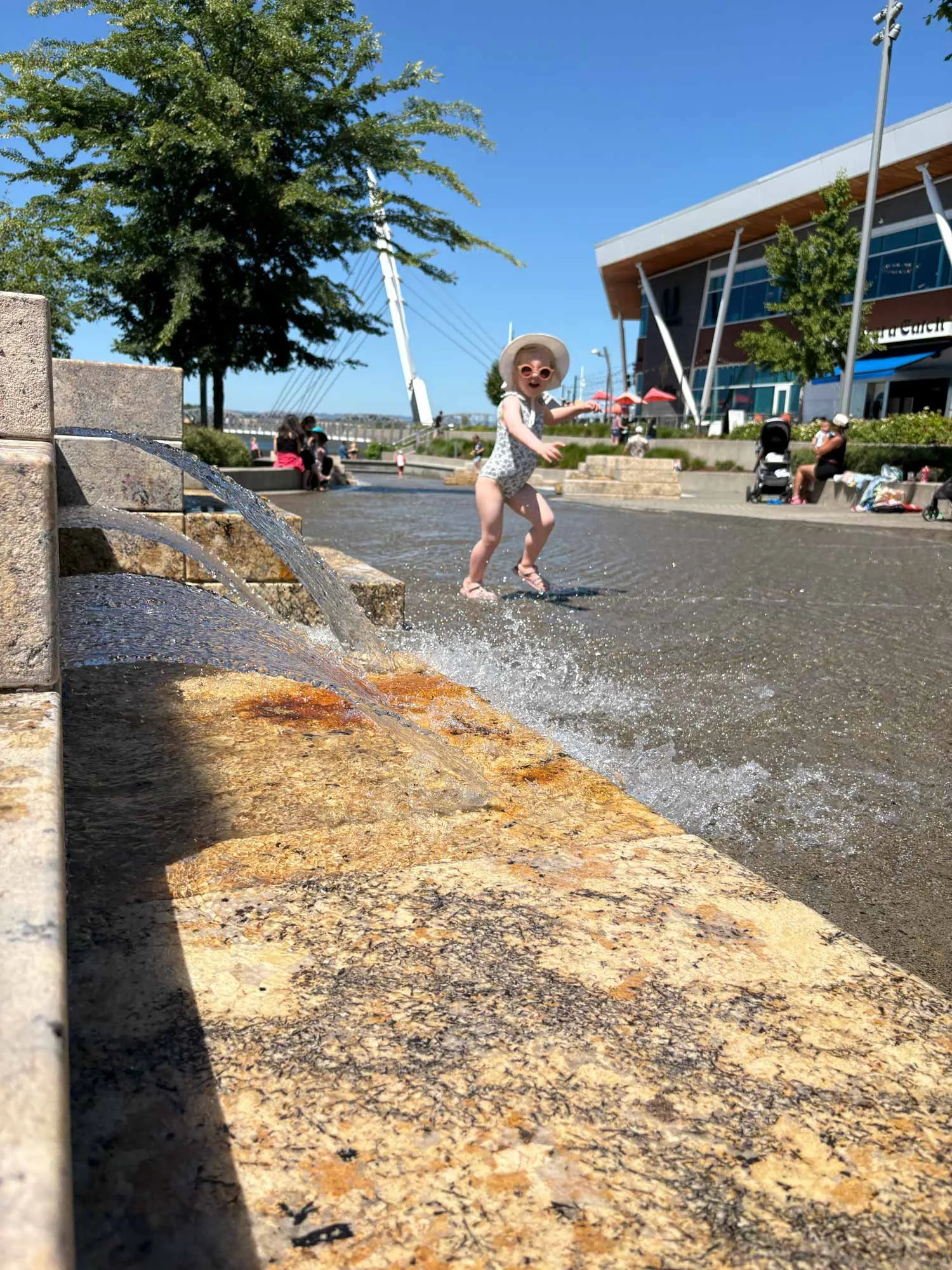 A young girl wearing a swimsuit, sunhat, and sunglasses playing in a shallow water feature at a park on a sunny day, with people sitting on benches and shopping area in the background.
