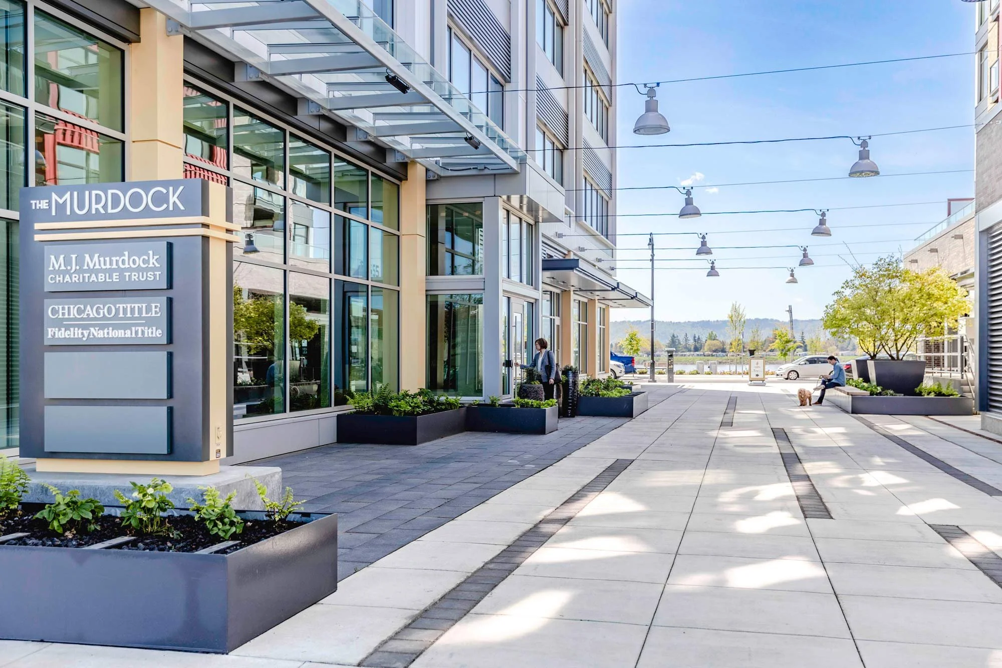 Modern commercial building with a sign for M. J. Murdock Charitable Trust and Chicago Title. People are walking and sitting outside on a sidewalk with planters, trees, and streetlights under a clear blue sky.