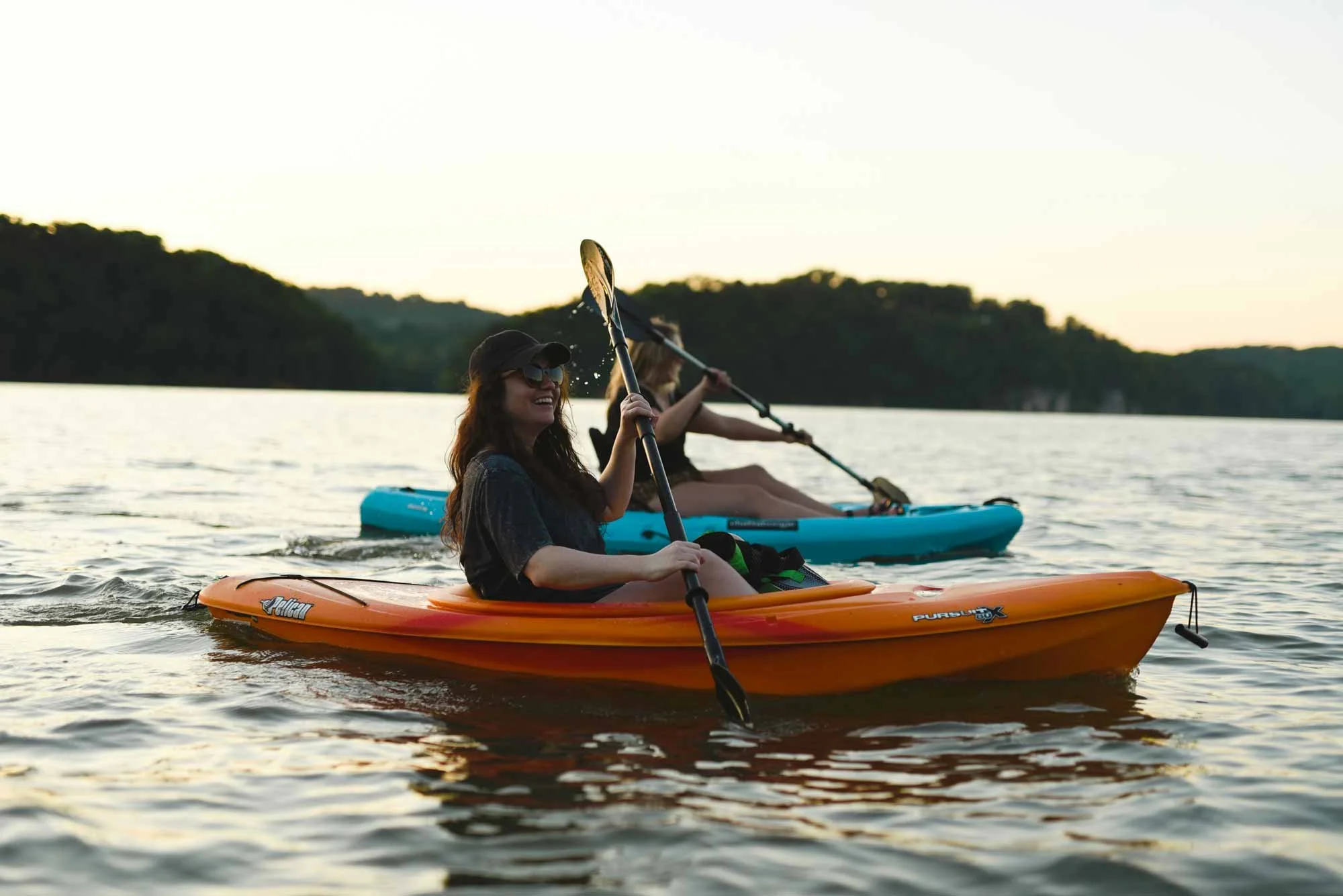 Two women kayaking on a lake during sunset, one in an orange kayak wearing sunglasses and a cap, the other in a blue kayak.