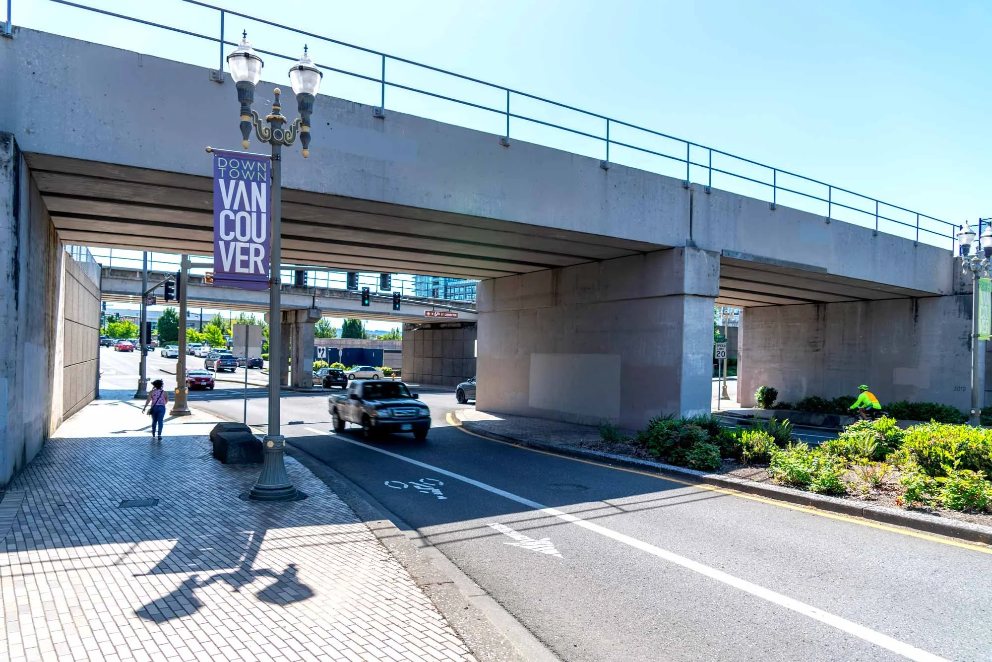 Underpass with a concrete bridge, street lamps, a pedestrian and vehicles, a cyclist, and road signs.