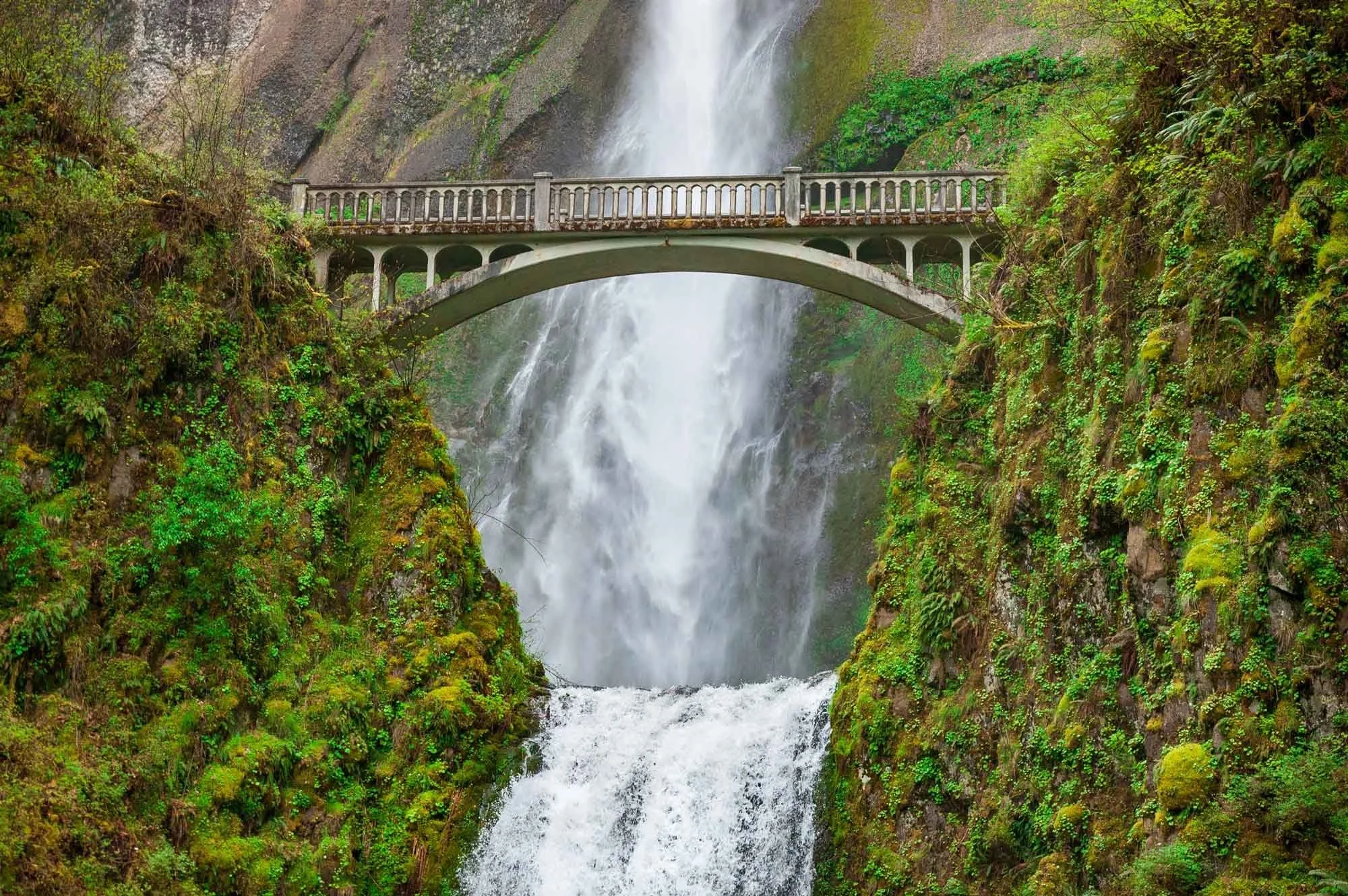 A waterfall cascading down a rocky cliffside surrounded by green vegetation, with a concrete bridge spanning across the river below.