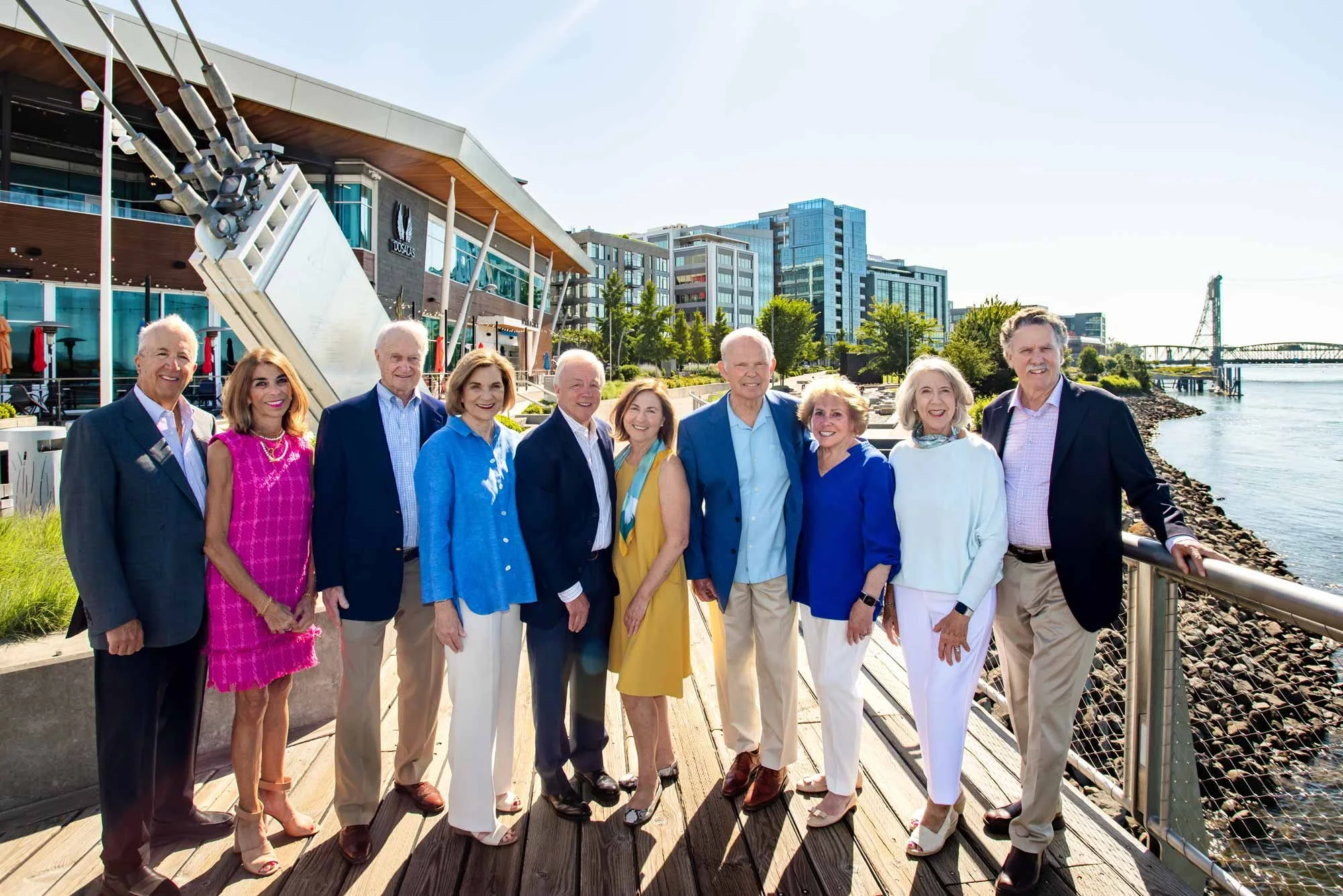 Group of eleven elderly people, six women and five men, standing on a wooden boardwalk by a waterway, with modern buildings and trees in the background. They are dressed in smart casual and business attire, smiling for the camera on a sunny day.