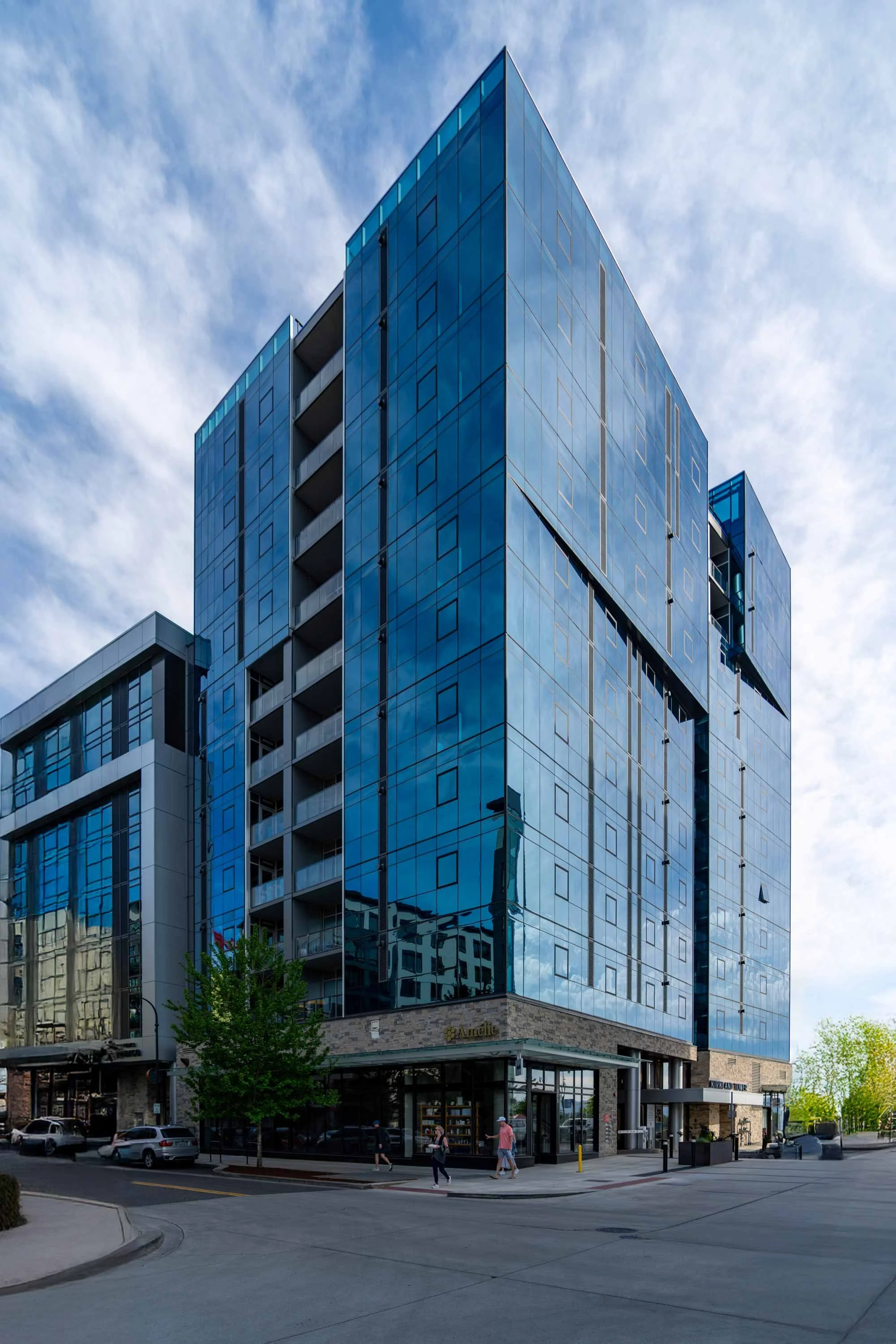 A modern glass high-rise building with retail shops at street level, a few pedestrians walking, parked cars, and trees in an urban area during daytime.