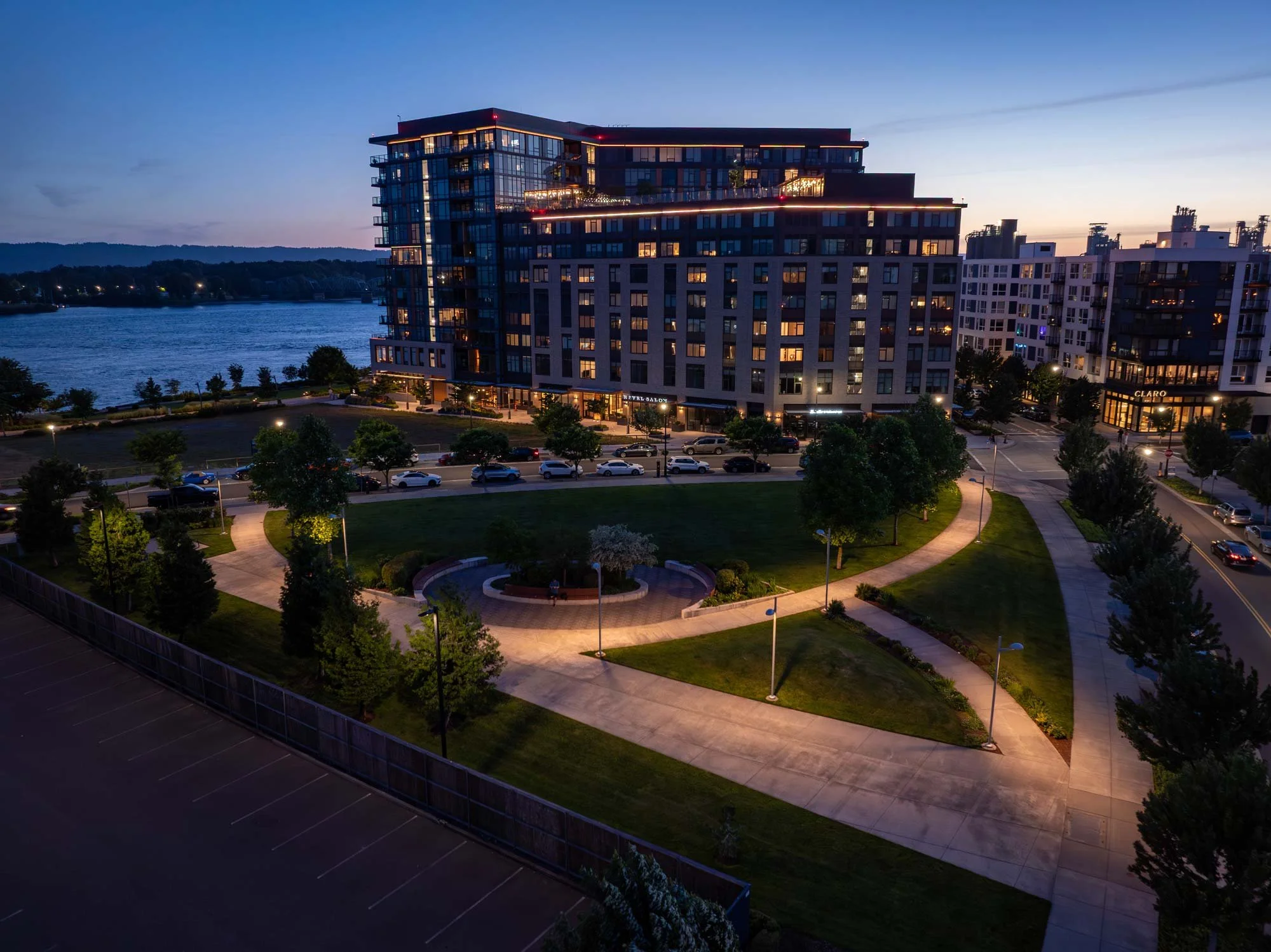 A modern multi-story residential building along a waterfront, nighttime with lit windows and outdoor lighting, surrounded by a park with walking paths, trees, and parked cars.