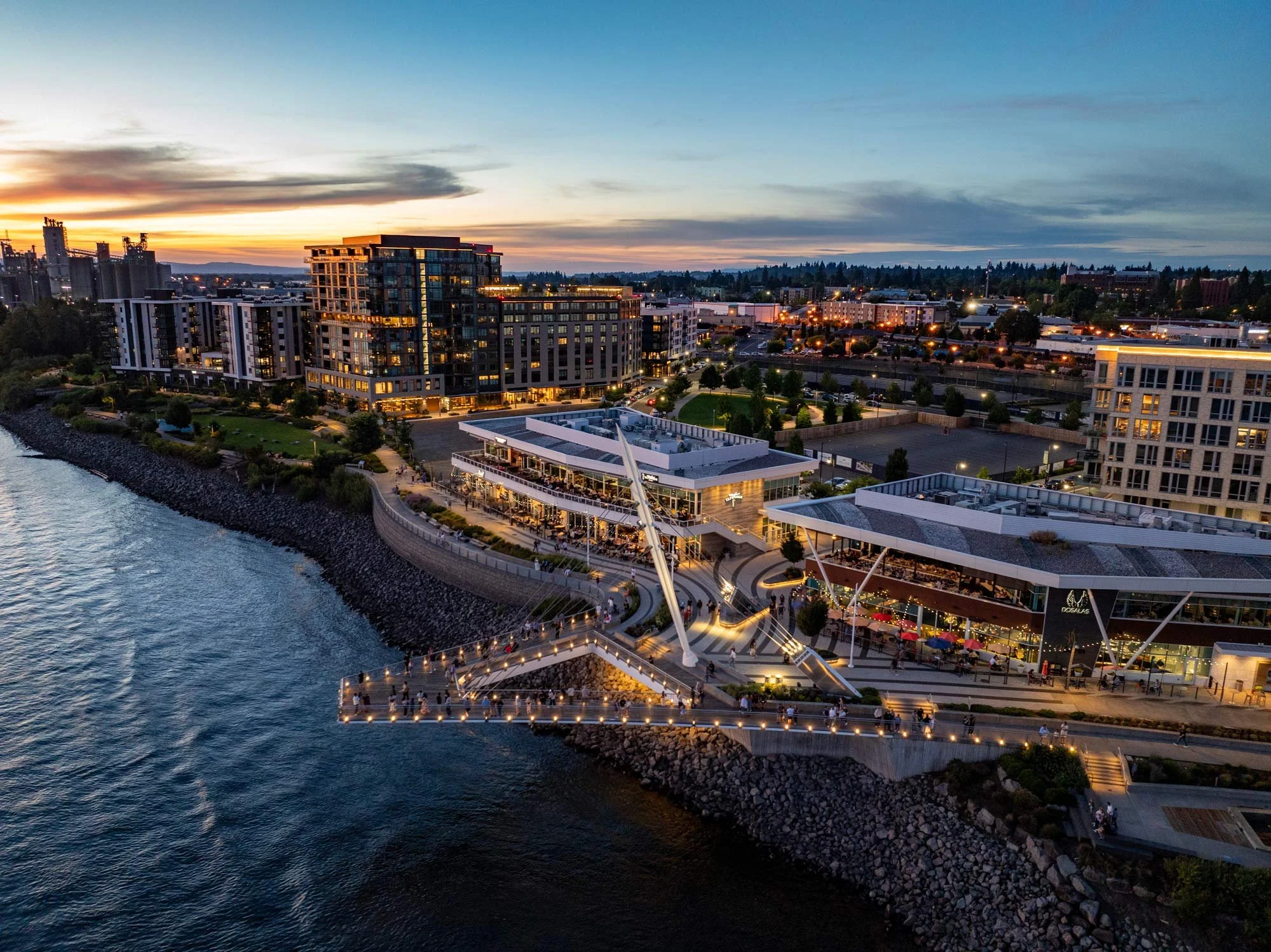 Aerial view of a waterfront cityscape at sunset. Shows modern high-rise buildings, a pedestrian bridge over water, and streets with illuminated streetlights.