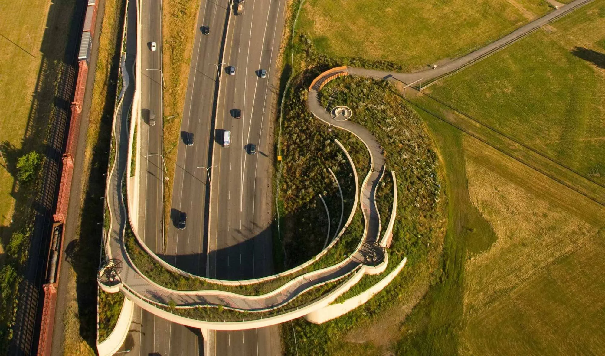 Aerial view of a winding walkway circling a landscaped area next to a highway with multiple cars.