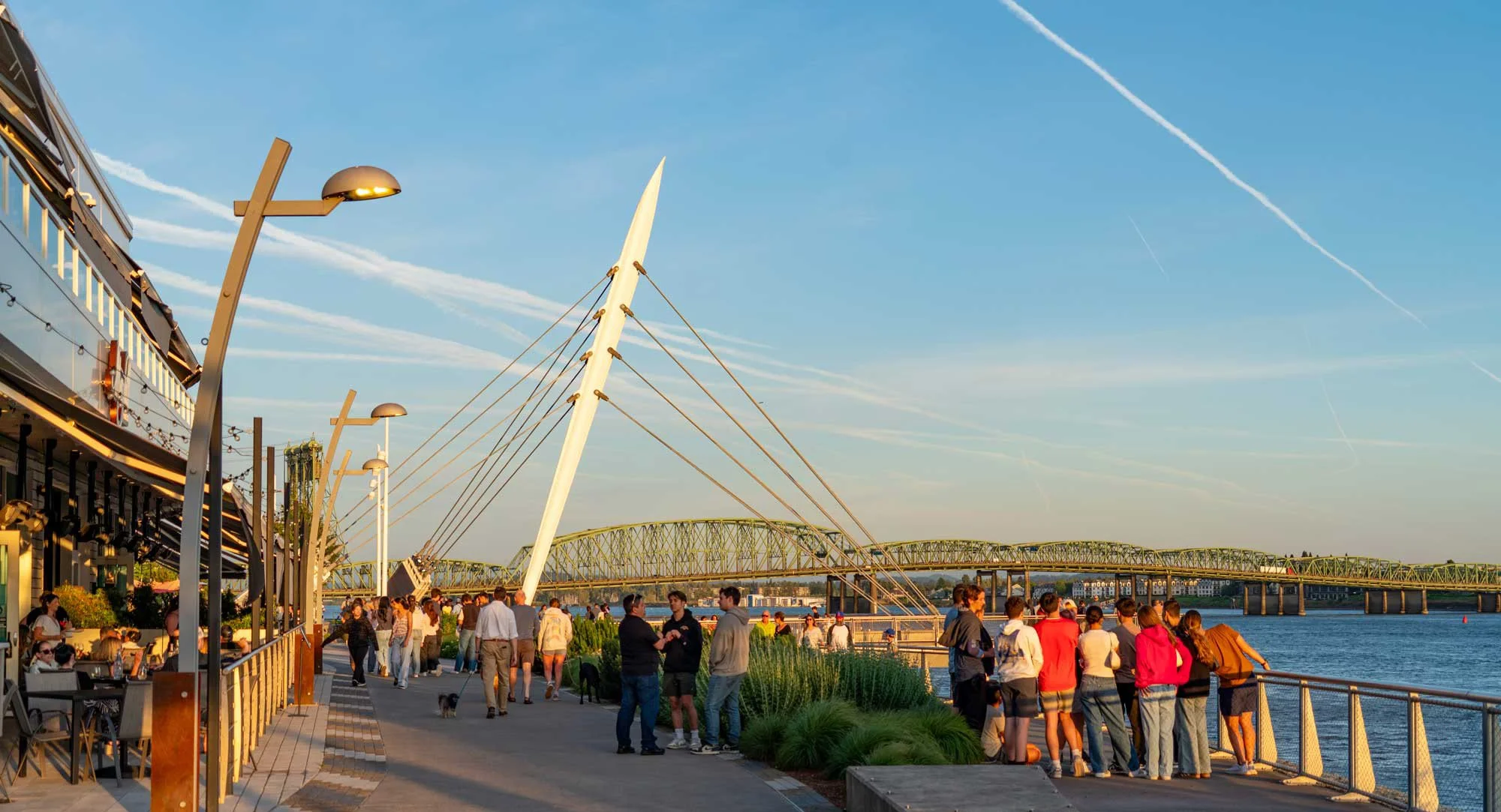 People walking and gathering along a riverside promenade with a modern bridge and blue sky in the background.