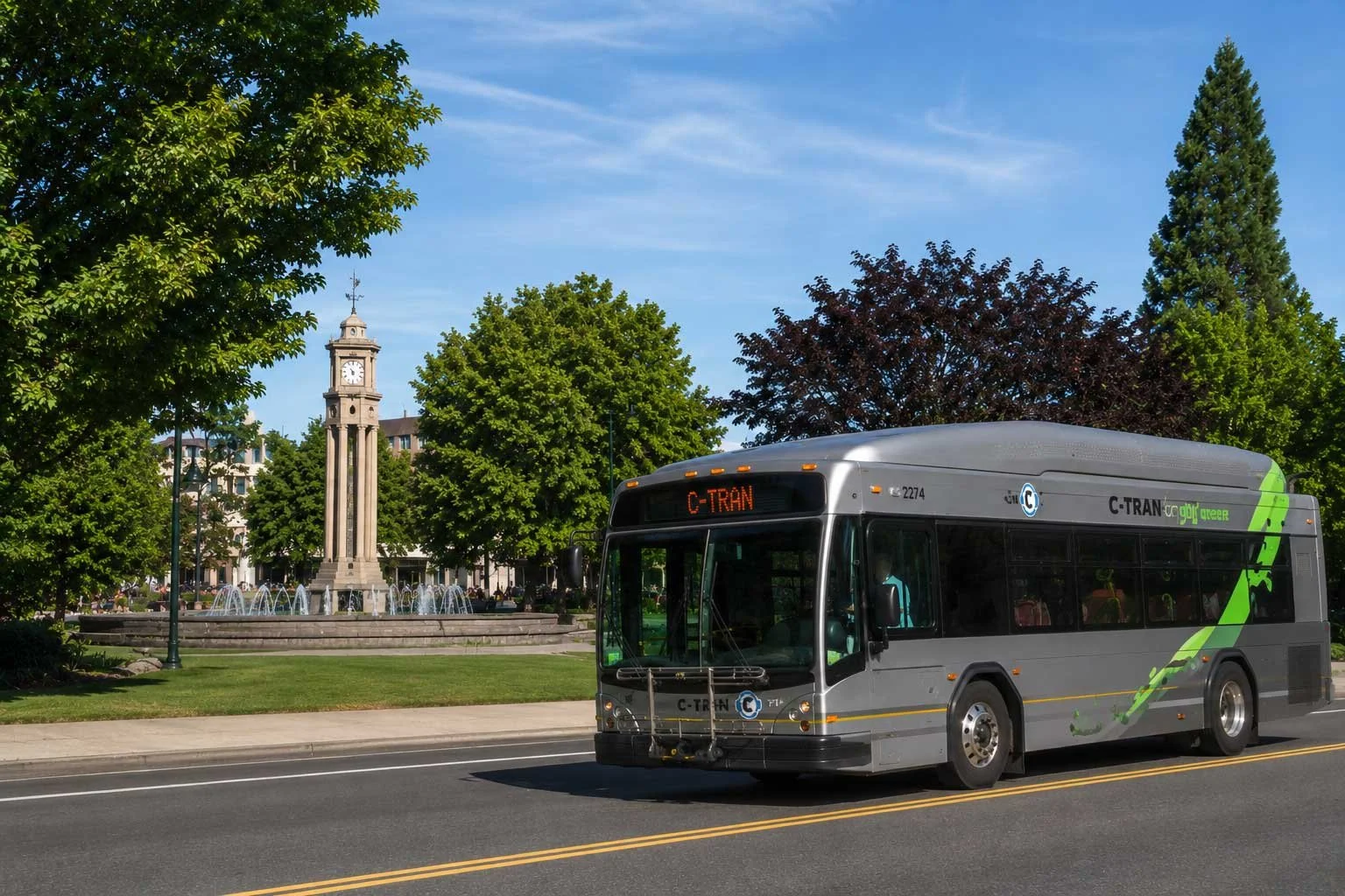 A city street with a silver C-Tran bus driving on the road. There is a park with green trees, a fountain, and a clock tower in the background under a blue sky.
