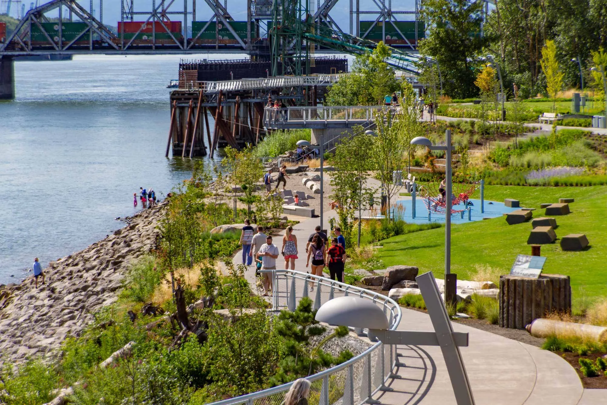 Park along a waterfront with people walking, sitting, and playing on playground equipment. A bridge and cargo ships are visible across the water, with greenery and trees lining the park.