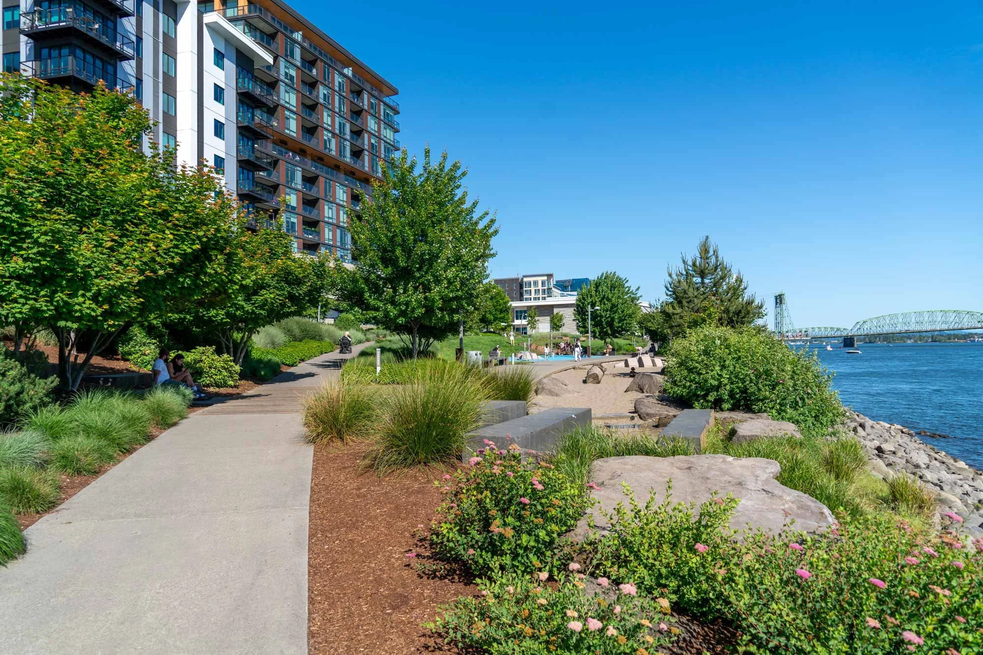 A riverside park with a paved walking path, green trees, flowering bushes, and benches, alongside a river with boats, and a bridge in the distance under a clear blue sky.