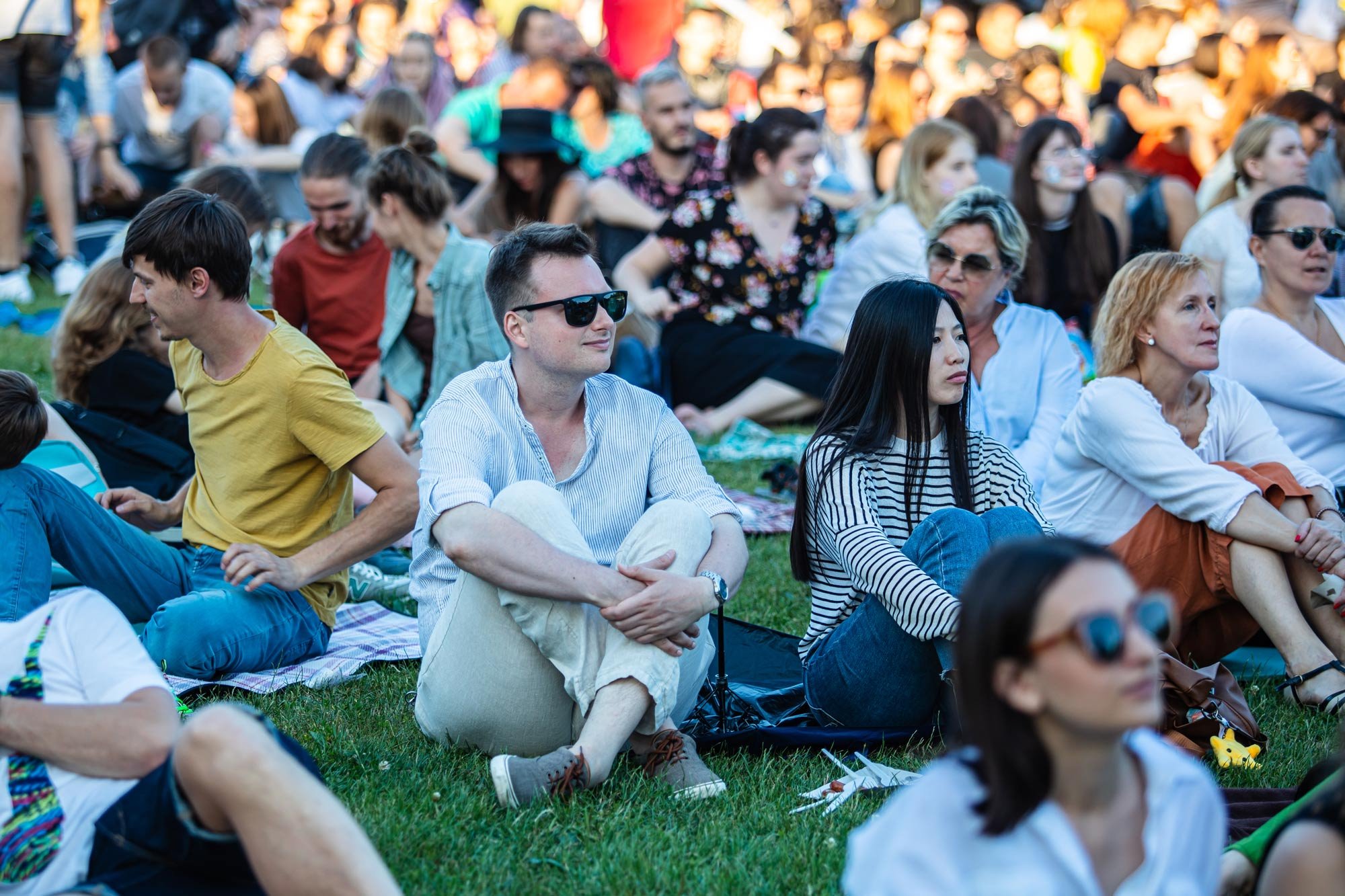 People sitting on grass at an outdoor event, some are watching or listening attentively, others are talking or relaxing, during the daytime with sunlight.