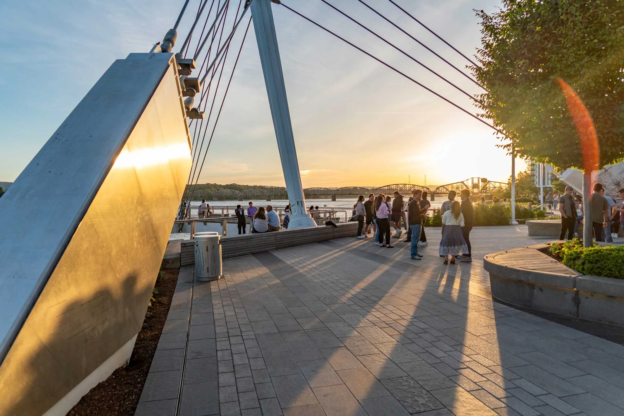 People gathering near a waterfront during sunset, with a bridge in the background, trees, and an architectural installation reflecting the sunlight.