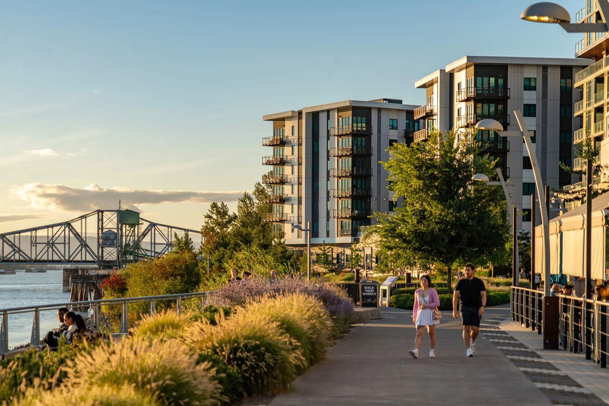 People walking along a riverside promenade with modern apartment buildings, trees, and a bridge in the background during late afternoon sunlight.