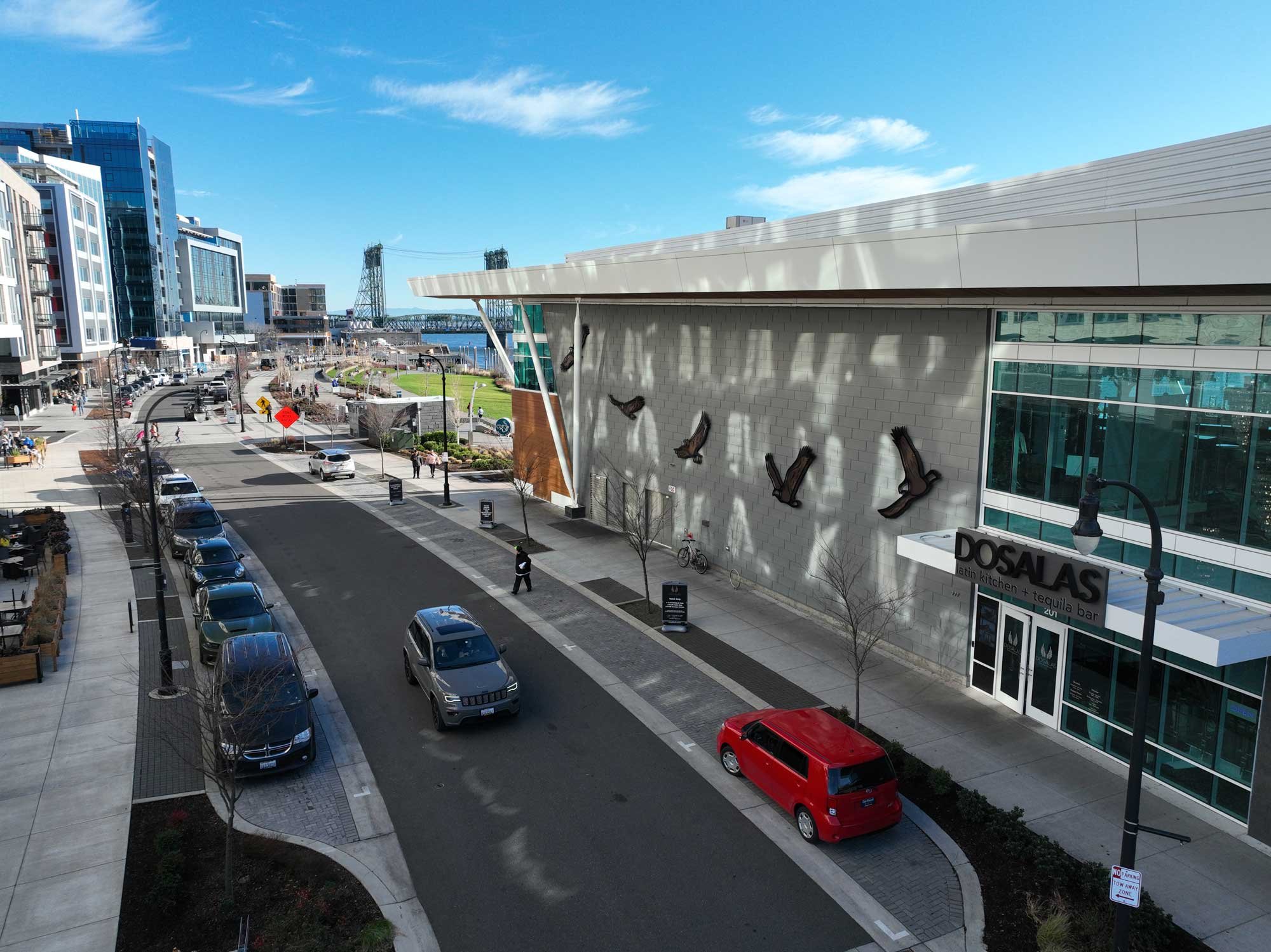 Urban street scene with parked cars, modern buildings, a waterway in the background, and a bridge. Clear blue sky with some clouds.