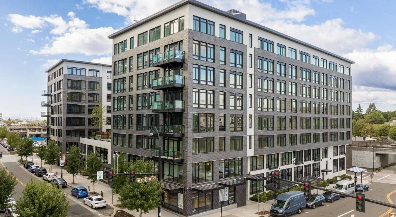 Modern multi-story residential building with large windows and glass balconies, located at the corner of a street with parked cars and trees, under a partly cloudy sky.
