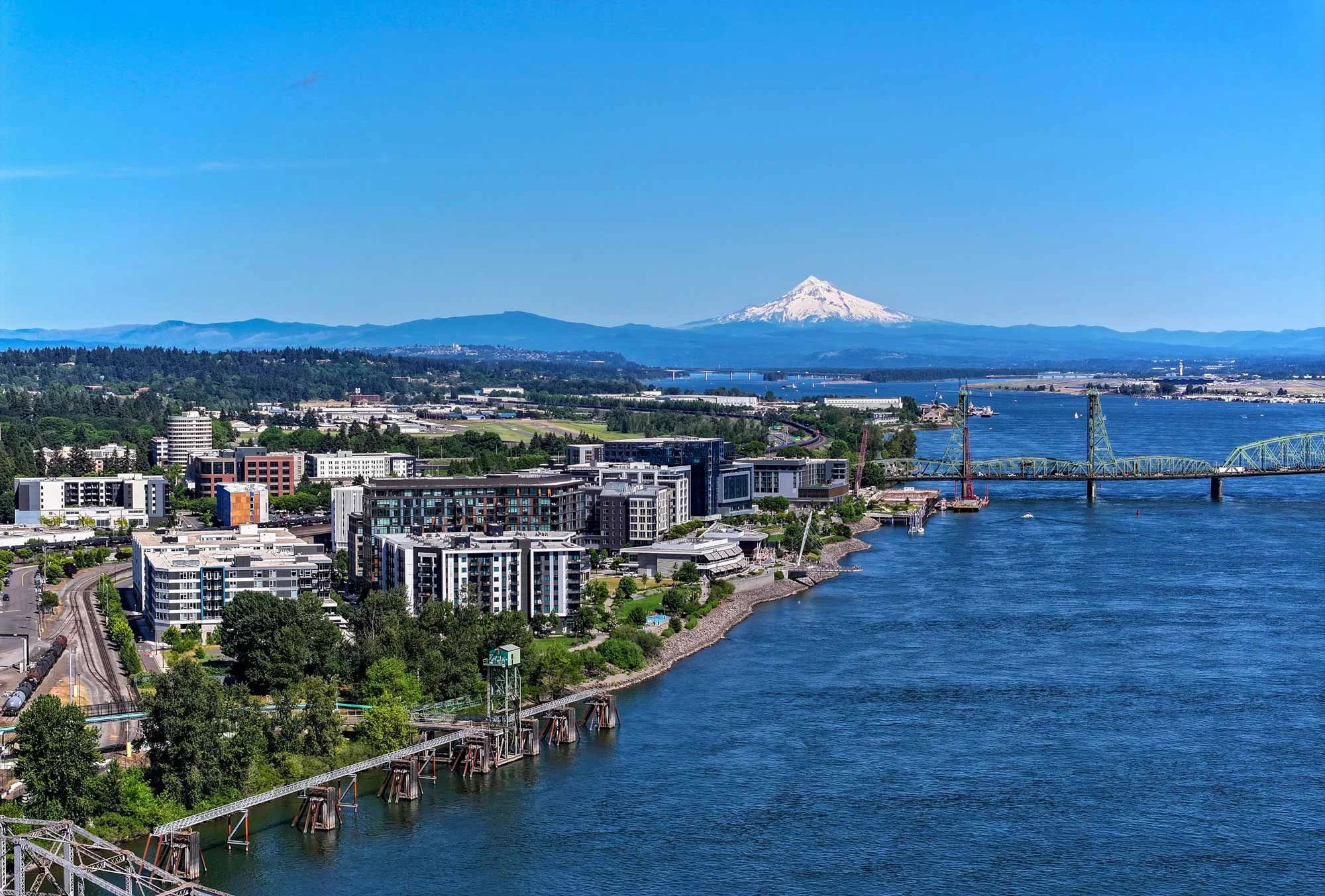 A cityscape with modern buildings along a river, a bridge crossing the water, and a snow-capped mountain in the background under a clear blue sky.