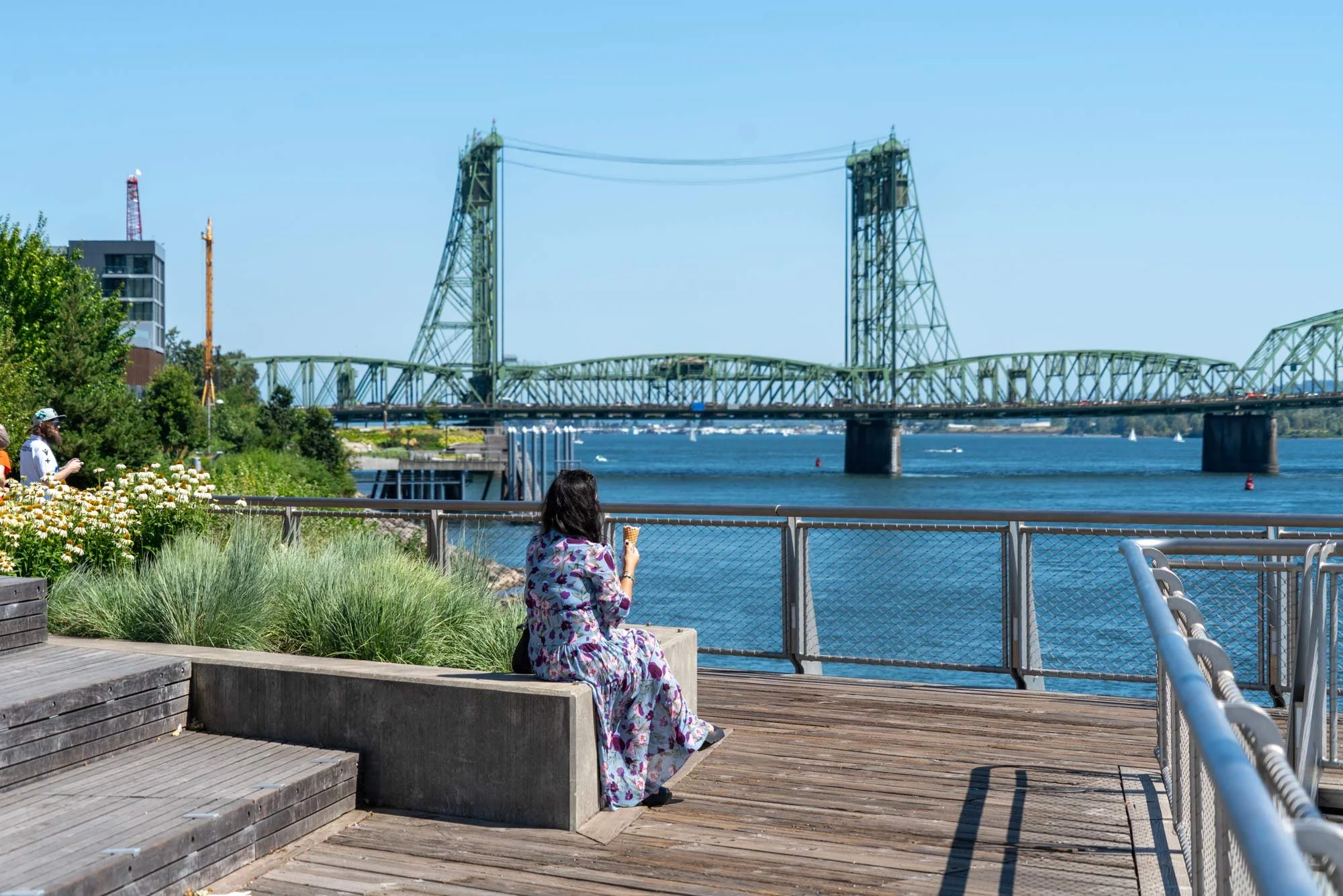 A woman in a floral dress sitting on a concrete seat by a waterfront, with a woman in a white hat and a man on the left side, and a green bridge over the water in the background under a clear blue sky.