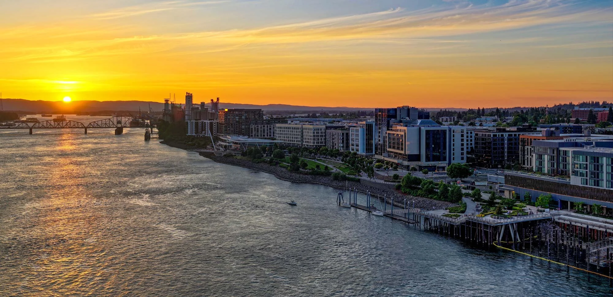 Sunset over a city waterfront with modern buildings, a bridge, and hills in the background.