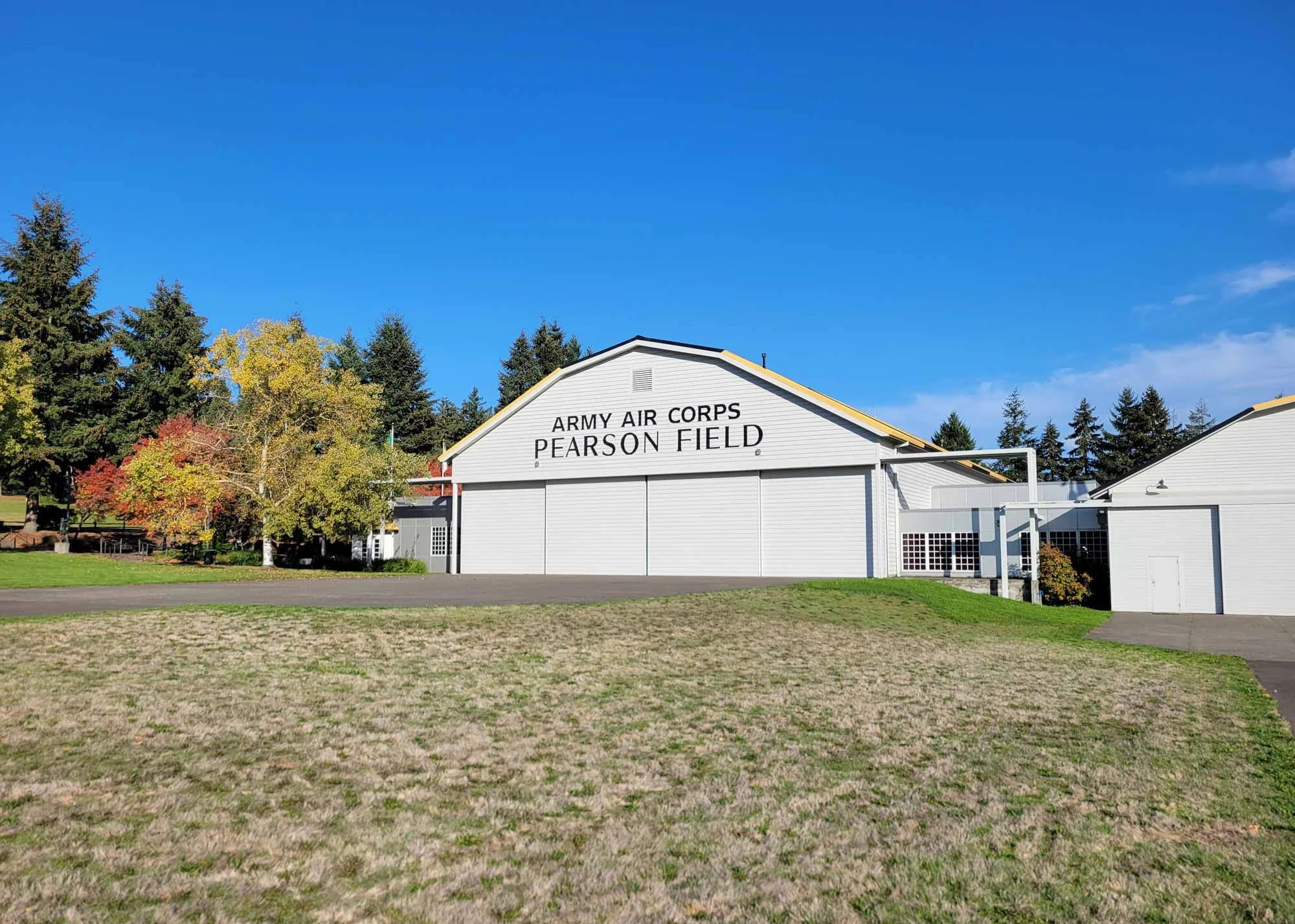 A white building with a sign that reads 'ARMY AIR CORPS PEARSON FIELD' is surrounded by trees with fall foliage, under a clear blue sky.